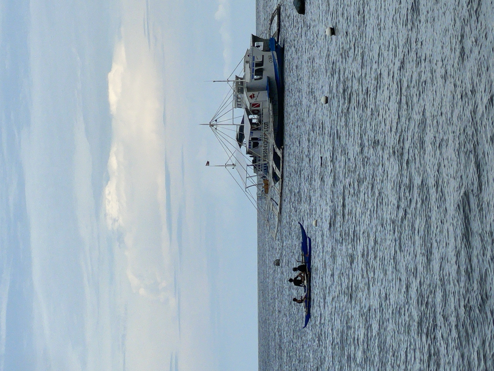 A scene captures the ocean off the coast of Dauin, Philippines, under a partly cloudy sky. In the foreground, a small blue outrigger canoe with three occupants is visible. Two individuals are seated towards the rear, while another sits closer to the front. The canoe is in motion, with ripples emanating from its hull.
Further in the distance, a larger, white and blue passenger ferry, possibly a dive boat, is anchored. The ferry has multiple decks, a prominent superstructure, and an outrigger design characteristic of Filipino vessels. A red flag is visible on a mast. Text on the side of the ferry reads "www.sea-explorers.com". Several white buoys are scattered across the water's surface. The water itself is a deep blue-gray, with moderate ripples and waves. The horizon is clearly defined, separating the sea from the sky.