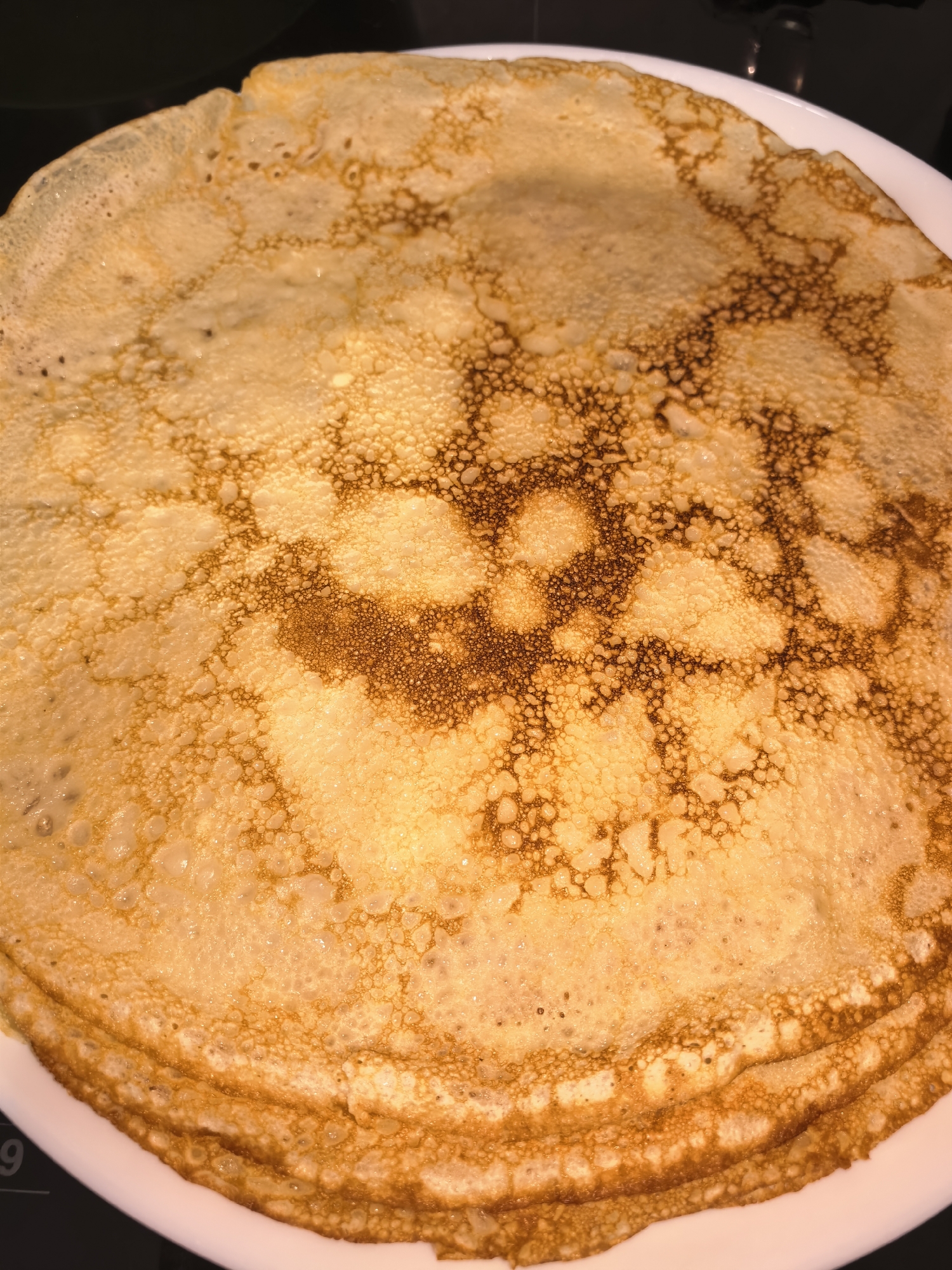 The image displays a close-up, top-down view of a stack of golden-brown crepes or thin pancakes. The crepes are round and rest on a white plate, which is partially visible along the bottom and sides of the frame. The surface of the topmost crepe is covered in intricate patterns of lighter golden and darker amber-brown, suggesting areas of varying crispiness and doneness from cooking. Numerous small bubbles and porous textures are visible across the surface, characteristic of crepes. Several layers of the thin pancakes are visible, creating a soft stack. The lighting is warm, enhancing the inviting, rich color of the food. The background is dark and out of focus, ensuring the focus remains entirely on the crepes. A faint, blurry numeric digit, possibly a "9," is subtly visible in the bottom-left corner from the hints, which could be part of a kitchen appliance display. No specific activity is depicted, but the crepes appear freshly made and ready for consumption.