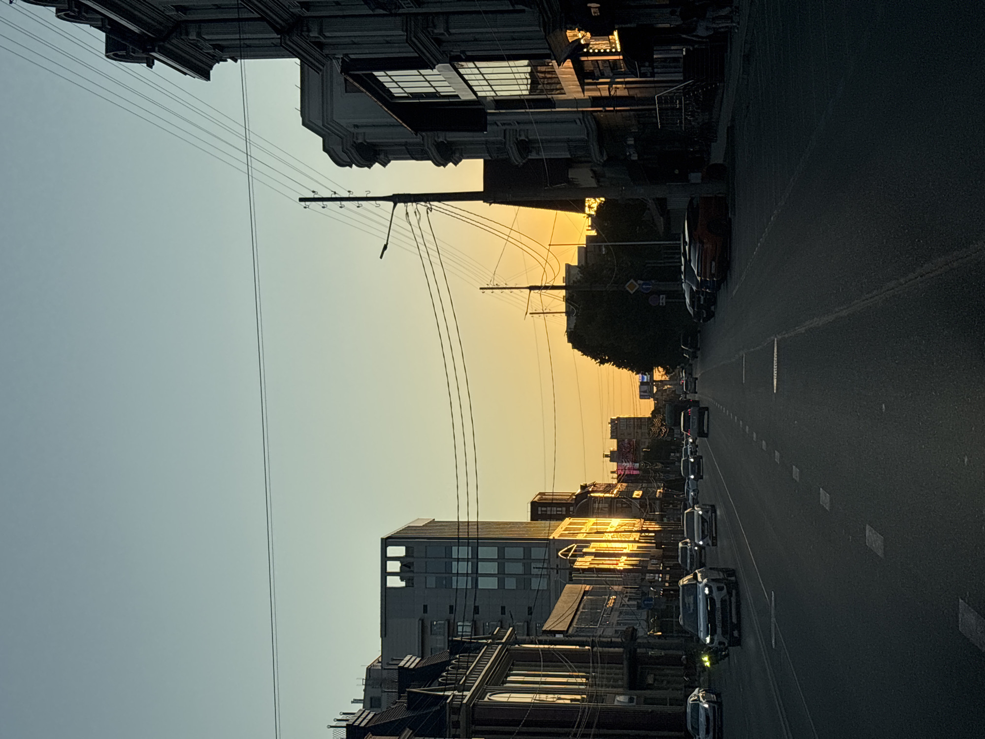 The image depicts a street scene in Krasnodar, Russia, during sunset. The sky is a gradient of pale blue to warm orange, indicating the low position of the sun. The street is paved and marked with dashed white lines, suggesting it is a multi-lane road. Several cars are visible on the road, driving in both directions. On the left side of the street, there are buildings with ornate architecture, some reflecting the warm sunset light. Further down the street on the left, a modern, taller building with a grid-like facade stands out. On the right side of the street, there are also buildings, some with commercial storefronts that are illuminated. Power lines and utility poles are prominent throughout the scene, crisscrossing the sky. Trees and foliage are visible in the background, particularly on the right side, partially obscuring some buildings and adding to the urban landscape. The overall impression is an active city street at dusk, bathed in the soft glow of the setting sun.