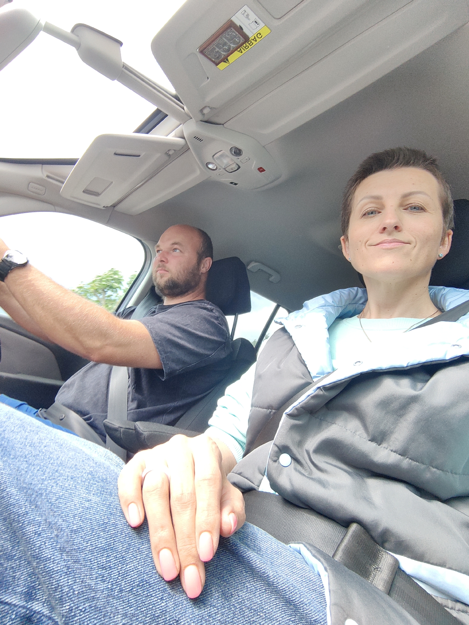 In the foreground, a person's hands, with polished pink fingernails, rest on their lap covered in blue jeans. Next to them, a woman with short brown hair and a light blue top underneath a grey and light blue puffer jacket is visible, looking towards the camera with a slight smile. Her seatbelt is fastened. In the background, a man with a beard and a dark t-shirt is driving, also wearing a seatbelt. The scene takes place inside a car with a sunroof open. The car appears to be on a road, with trees visible through the window. The lighting suggests it is daytime. The car is likely in the vicinity of Фаніпальскі сельскі Савет, Belarus, given the user's prompt specifying this location.