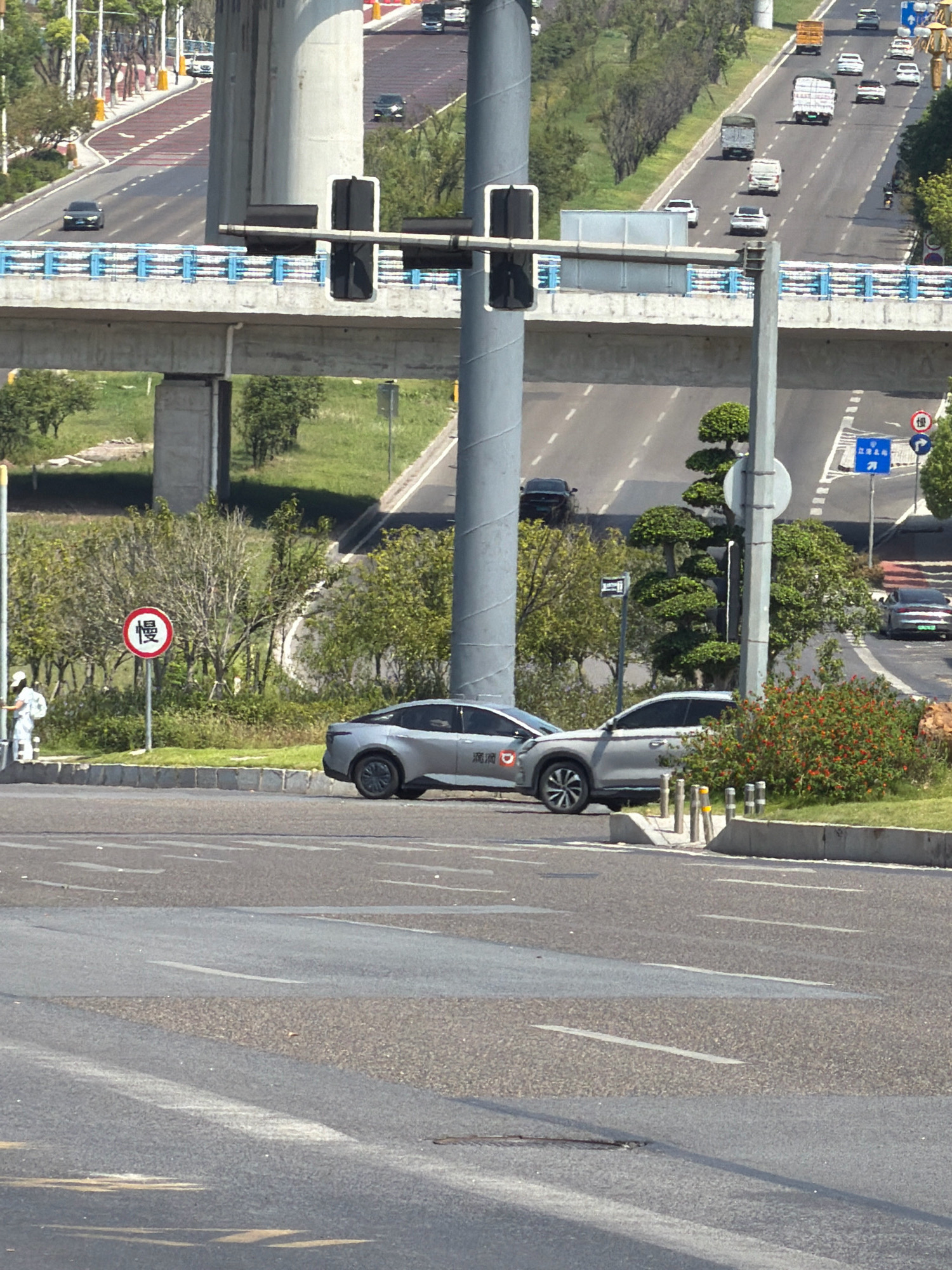 The image captures a street scene in Jiangjin, China, featuring a multi-level road structure with overhead bridges and elevated roadways. In the foreground, a grey sedan is parked at the side of the road, partially visible on the right. To the left of the car, a person in white protective clothing is standing near a red and white circular sign with Chinese characters that appear to translate to "slow." The road surface is asphalt, marked with white lines. The background shows other vehicles on the elevated roadways, trees, and supporting structures of the bridges. The sky is clear and bright, suggesting a daytime setting.