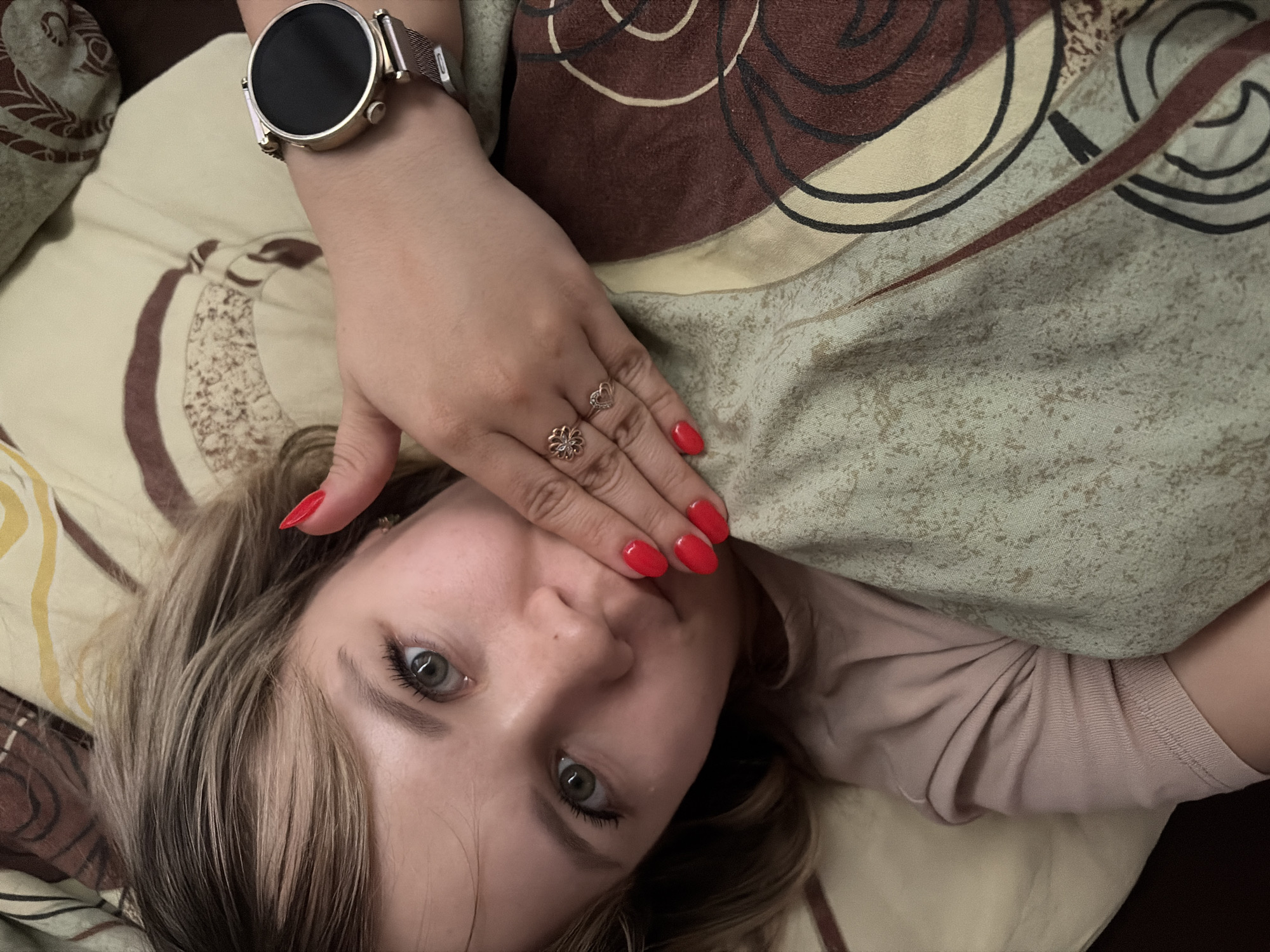 A close-up, top-down shot shows a young woman lying down, partially covered by a patterned blanket. She is looking directly at the camera with her left hand holding up her right hand which covers part of her mouth and nose.
The woman has light brown/blonde streaked hair, light green/hazel eyes with visible eyelashes, and a neutral expression. Her fingernails are painted a vibrant red. On her left wrist, she wears a gold-toned smartwatch with a black screen and a metallic mesh strap. Her right hand, which is covering her face, features two rings: one on her ring finger, shaped like a flower, and another on her middle finger, shaped like a heart. She appears to be wearing a light pink or beige long-sleeved top, visible on her right arm beneath the blanket.
The bedding consists of a blanket or duvet with an abstract pattern of brown, beige, and black swirling lines, mixed with areas of a speckled light green/beige texture. Another striped piece of bedding in yellow and brown is visible on the left side of the frame. The lighting is soft and even, suggesting an indoor setting, likely a bedroom. No visible text is present in the image.