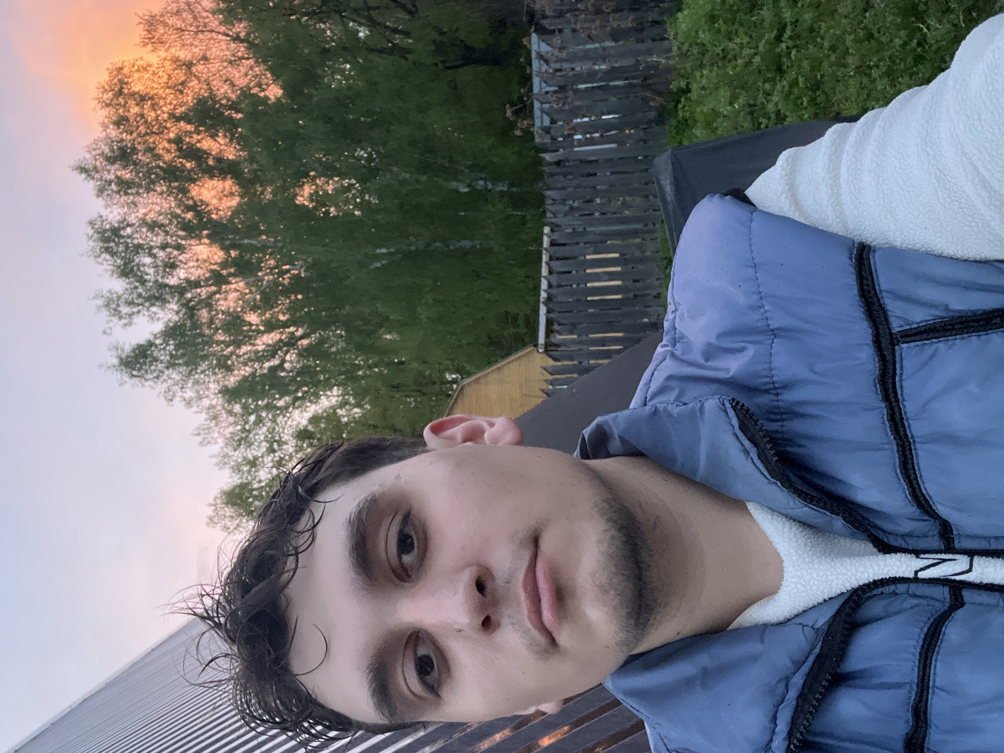 A selfie taken outdoors shows a young man with dark hair and stubble wearing a light blue puffy vest over a white sweater. He appears to be lying down, looking directly at the camera. Behind him, the background includes a wooden fence, a building with a yellow-painted section, a tree with green leaves, and a sky with a pink-orange gradient suggesting sunset or sunrise. The location appears to be near a building with a corrugated metal roof. Based on the prompt, the location may be Vasilyevo, Russia.