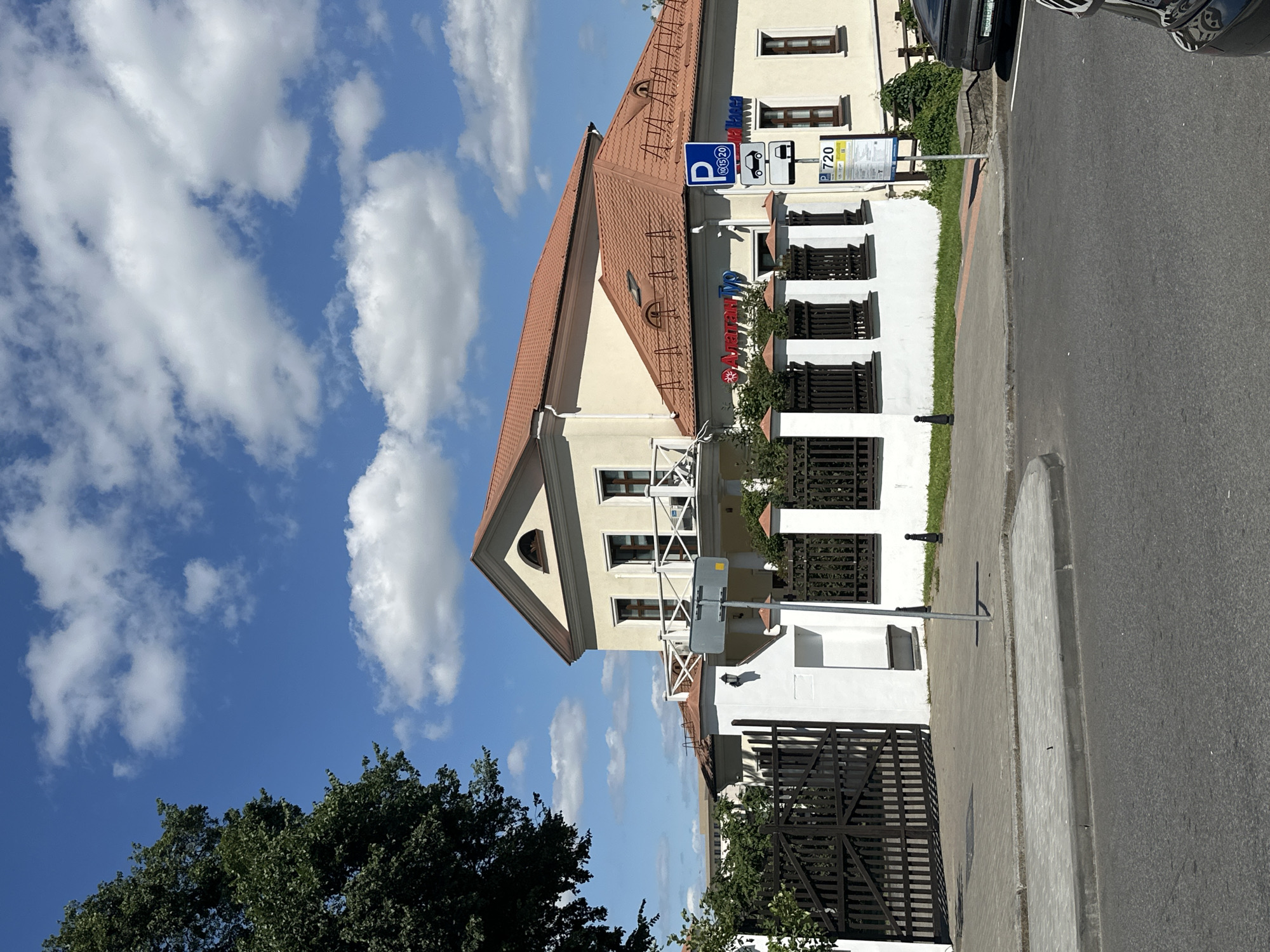 The image depicts a commercial building with a light yellow facade and a reddish-brown tiled roof, located in Minsk, Belarus. The building appears to be an office or retail space, with prominent signage including "Алатан Тур" (Alatan Tour) and "ОплатаМасса" (OplataMassa). A parking sign is visible to the right of the building, indicating paid parking. In front of the building is a dark wooden gate and a white fence, with a sidewalk and asphalt road in the foreground. A large tree with green foliage is on the left side of the image. The sky is blue with scattered white clouds. No people are visible in the image. The scene appears to be a sunny day.