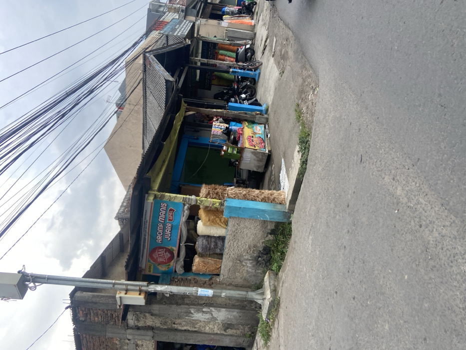 The image shows a street corner in Kecamatan Cimahi Selatan, Indonesia. A small shop with a blue awning and a sign that reads "Aroma Manis Juara Apel" is located on the corner. The shop is selling a variety of goods, including rolls of fabric and other merchandise. There are several motorcycles parked in front of the shop. The street is paved and there are power lines overhead. There are no people visible in the image. The image was captured during a cloudy day. The image was taken at the coordinates -6.93 latitude, 107.565 longitude.