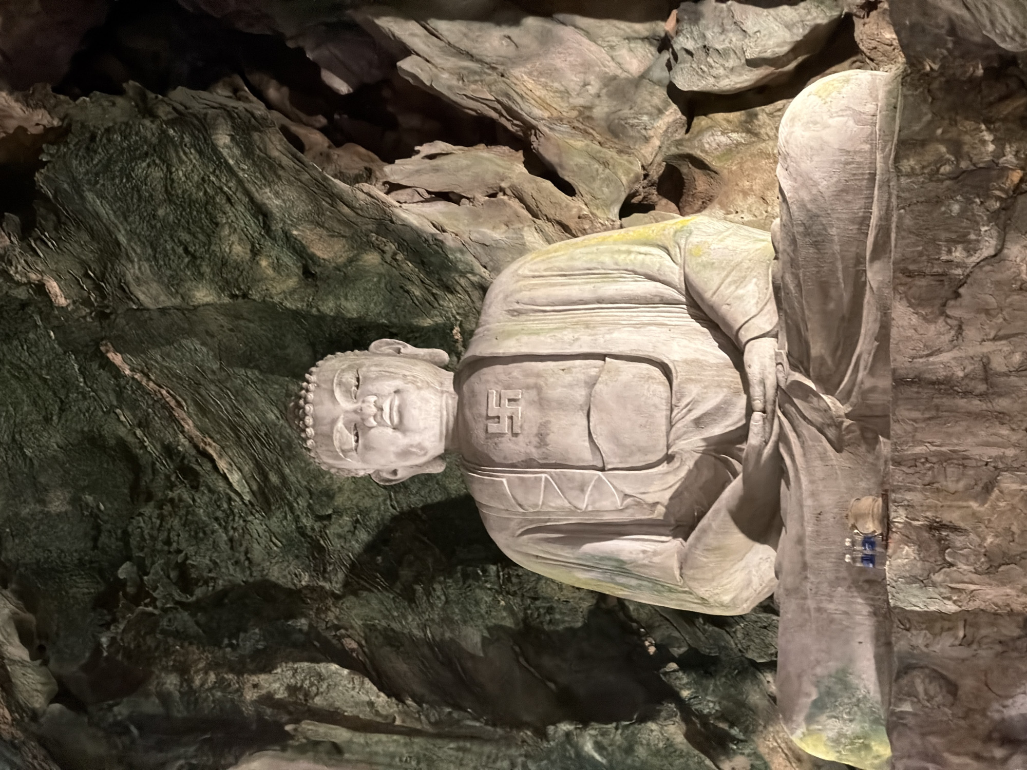 The image shows a seated Buddha statue carved into the rock walls of a cave. The statue is light grey in color and features a swastika symbol on its chest. The cave walls are textured with dark green and light brown tones. The statue's right leg is attached to the cave wall with small blue and white fixtures. This statue can be found in the Marble Mountains caves in the city of Đà Nẵng, Vietnam.