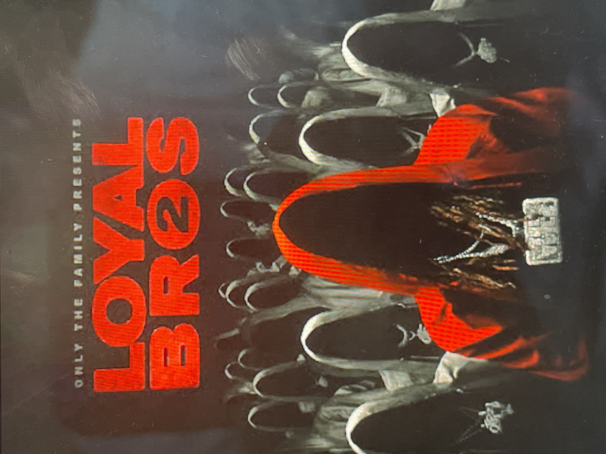 The image depicts what appears to be an album cover or promotional artwork, featuring a person from behind wearing a bright red hooded sweatshirt. The person has visible dreadlocks or braids and is adorned with a large, rectangular, silver-colored chain around their neck. The chain is embellished with what appear to be diamonds, and features text that is difficult to fully discern but resembles 'OTF'.
The background is dark with subtle textures, against which numerous pairs of black sneakers with white accents are arranged in receding rows, suggesting a crowd or a patterned graphic element. The overall lighting is dramatic, emphasizing the red hoodie and the metallic glint of the chain and shoes.
On the left side of the image, two blocks of text are visible, oriented vertically. The smaller text at the top reads 'ONLY THE FAMILY PRESENTS'. Below it, in large, bold red letters, the text 'LOYAL 2 BROS' is displayed, with '2' being a numeral. The aesthetic is characteristic of urban or hip-hop music branding. This media file is associated with Sofia, Bulgaria.