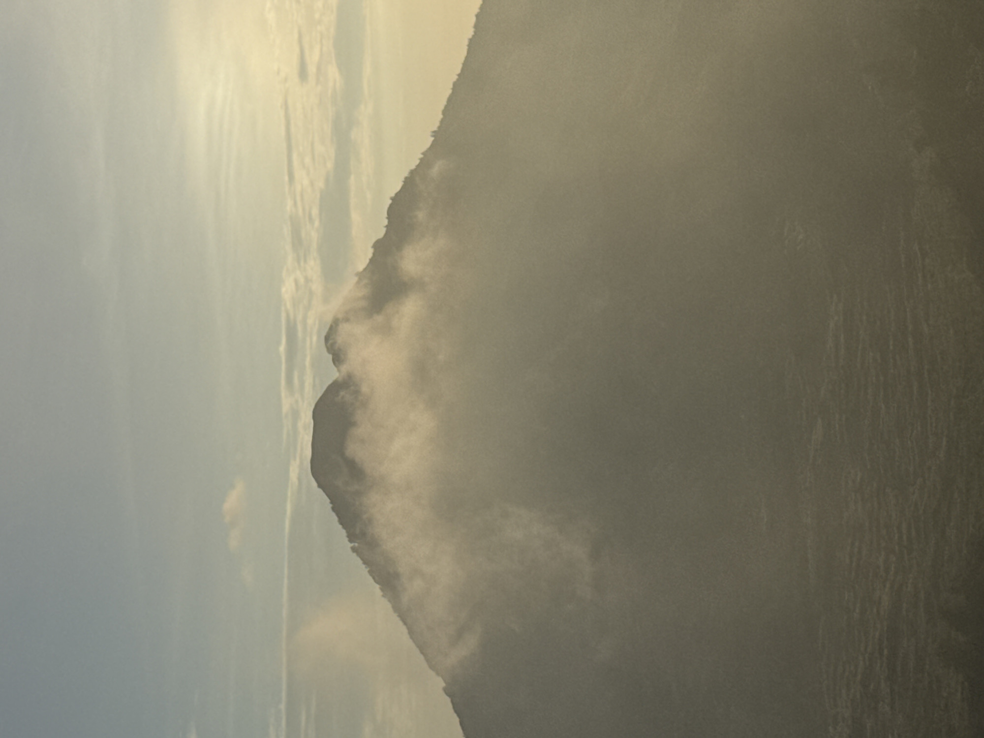 The image shows a hazy mountain landscape, partially obscured by mist or clouds. The scene is vertically oriented. A peak is prominent on the left, shrouded in white clouds or mist that partially obscure its form. To the right of the peak is the vast slope of a larger mountain, the exact details of which are obscured by dense haze. The sky to the left is a pale blue or grey, with wisps of cloud or vapor. The location is Kedisan, Indonesia.