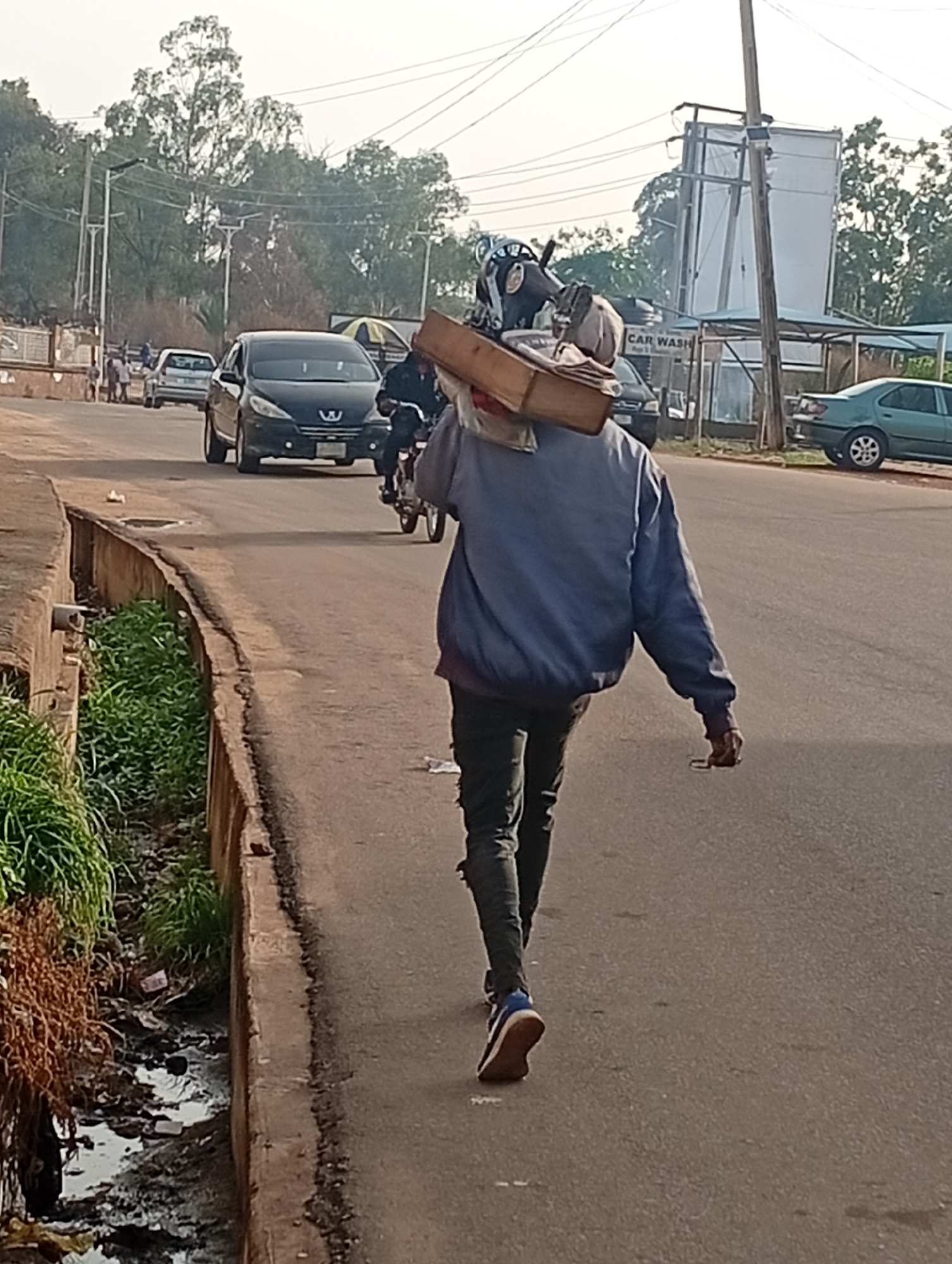 The image depicts a man walking away from the camera on a street in Gura Top, Nigeria. He is carrying a large, rectangular object on his shoulder, possibly containing various items including what appears to be a helmet. The man is wearing a blue jacket and dark pants, with blue and brown sneakers. He walks on the right side of a paved road, near a low concrete curb. To the left of the curb is a drainage ditch with some vegetation and litter. Traffic is visible on the road, including a black car and a motorcycle behind the man. In the background, there are buildings, trees, and utility poles. A sign that says "CAR WASH" is visible in the background.