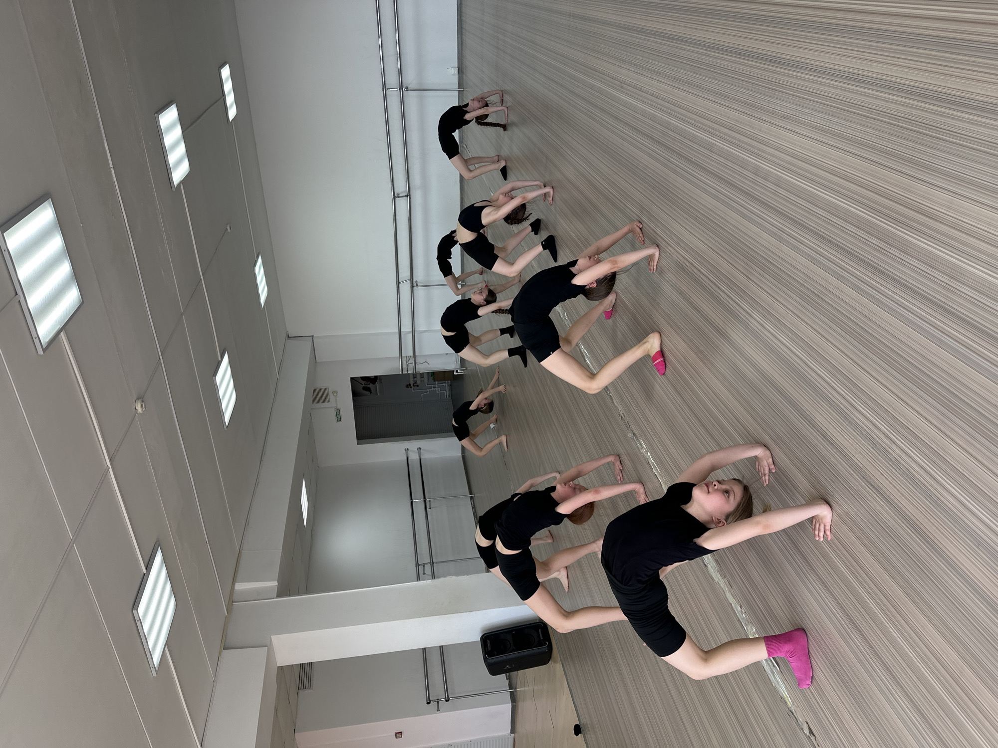 The image depicts a group of girls practicing a dance routine in a dance studio in Votkinsk, Russia. The studio has a polished, wooden floor with the girls arranged in a row, performing a backbend-like pose with arms extended. The girls are wearing dark athletic attire. The studio has bright, square overhead lights, a mirrored wall, and ballet barres. A black speaker sits near the front of the row.