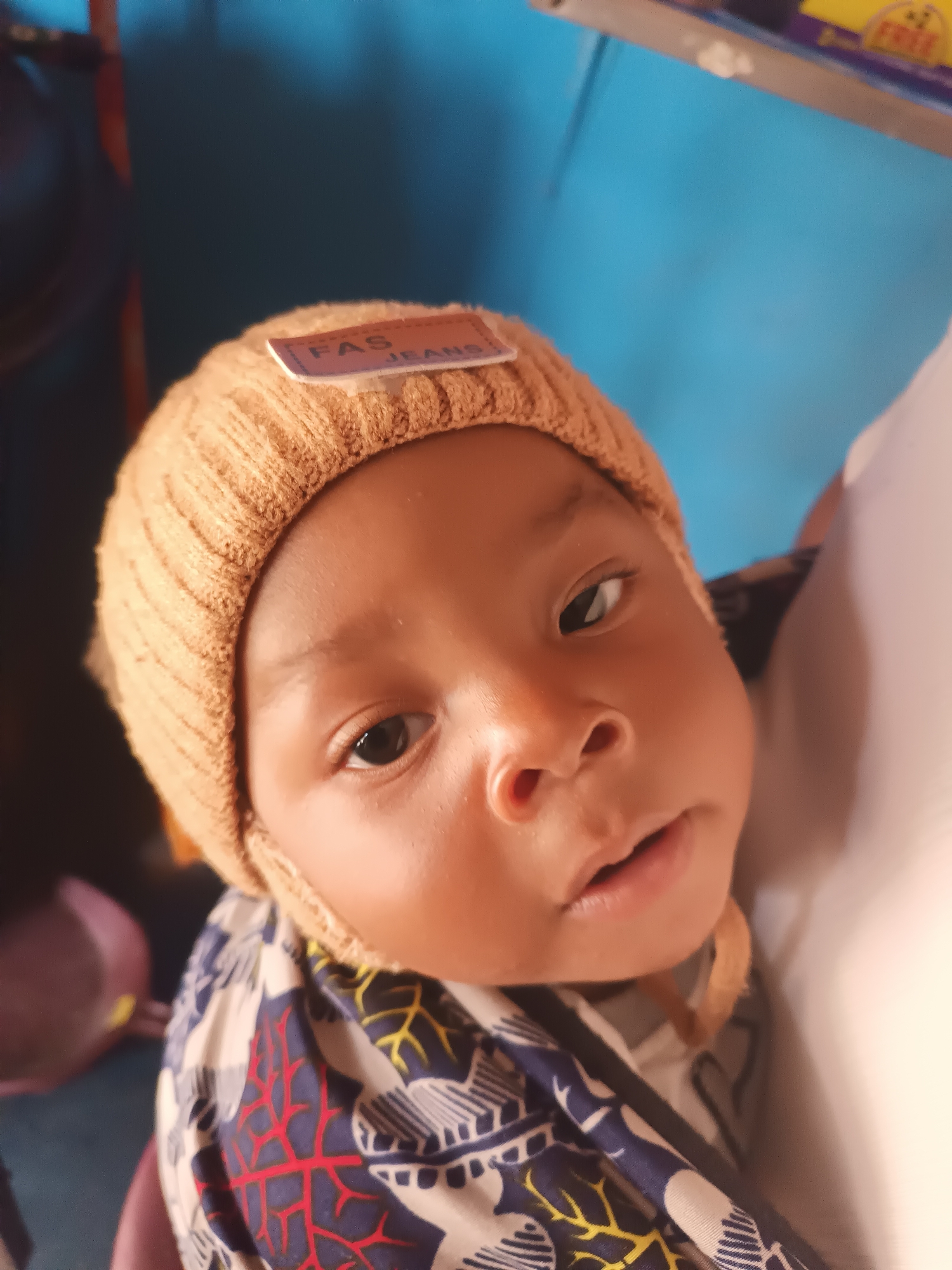 The media file is a close-up, portrait-style photograph of a baby's face. The baby is wearing a tan, ribbed knit beanie with a small rectangular tag that reads "FAS JEANS". The baby's skin tone is brown, and their eyes are dark. The background is predominantly a bright blue, suggesting an indoor setting, possibly a wall. To the right of the baby's head, a soft, white, out-of-focus area is visible, which could be a person holding the baby or a blanket. The baby appears to be looking slightly off-camera, with a neutral or slightly curious expression. The lighting suggests it might be daytime, casting soft shadows on the baby's face. The image is focused on the baby's upper body and face, highlighting their features and the hat. A brightly colored, patterned garment is visible around the baby's neck and shoulders, featuring blue, white, red, and yellow abstract designs. The overall impression is of a candid, intimate shot of a young child.
