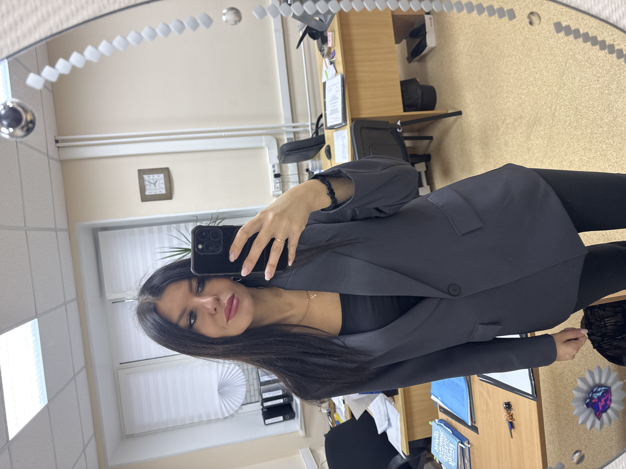 A woman is taking a selfie in a mirror. She has dark hair and is wearing a dark blazer and black top. The mirror is round with decorative trim.
The reflection shows an office environment. There are desks with office supplies, a chair, a clock on the wall, and blinds on the window. The location appears to be in a building in Chaikovsky, Russia.