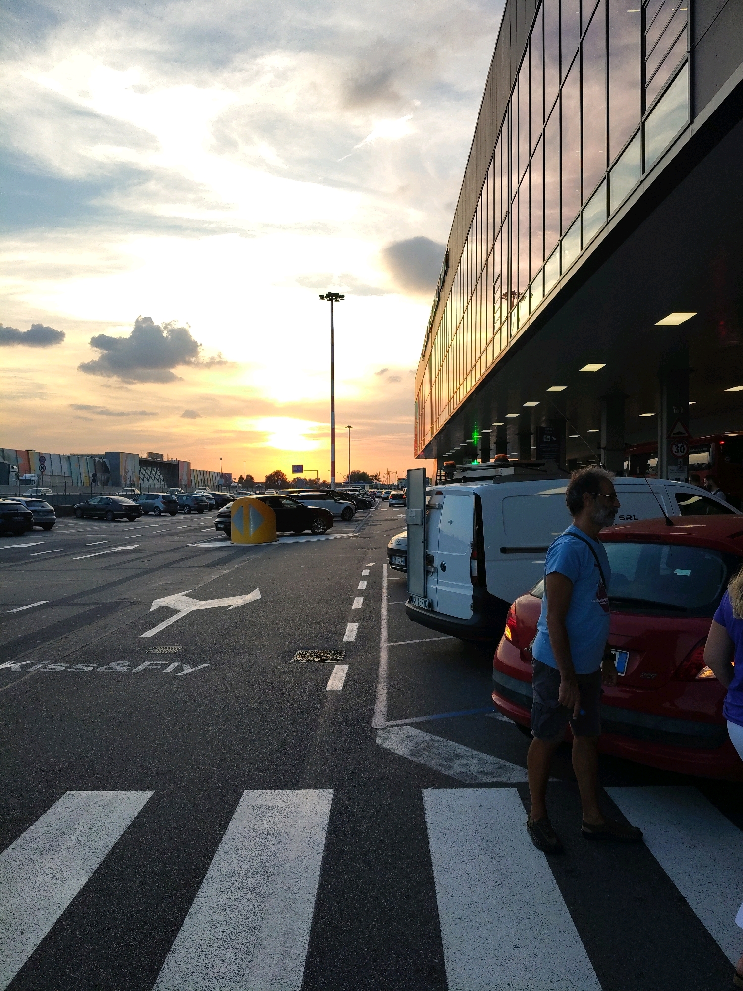 The image shows a man standing on a pedestrian crossing in front of a building with a reflective glass facade. He is wearing a blue t-shirt and grey shorts. A red car is parked to his right, and a white van is parked behind him. The sun is setting, casting a warm glow over the scene. There are other cars in the background, and a tall light pole. The ground is asphalt with white painted lines indicating pedestrian crossings and traffic lanes. The text "Kiss&Fly" is visible on the road. The scene appears to be an airport drop-off or pick-up area.