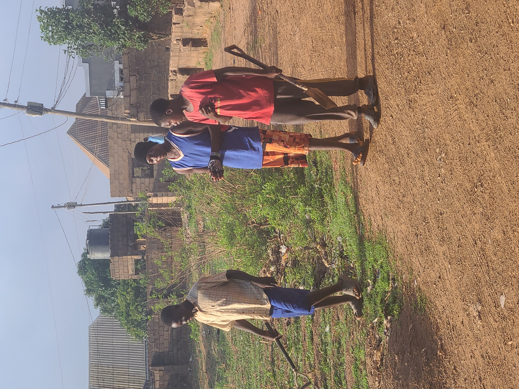 Three people are standing in Barakin Lerawi, Nigeria. The person closest to the viewer is wearing a light yellow button-down shirt and blue shorts and holding a shovel with the head of the shovel on the ground. The person behind him is wearing a blue tank top and orange and red patterned shorts. The last person is wearing a red t-shirt and dark pants and carrying a shovel on their shoulder. They are standing on a dirt path. A grassy area with weeds is to the left of the path. To the left of the grass are houses with cinder block walls. Behind the houses are a water tower and a utility pole.