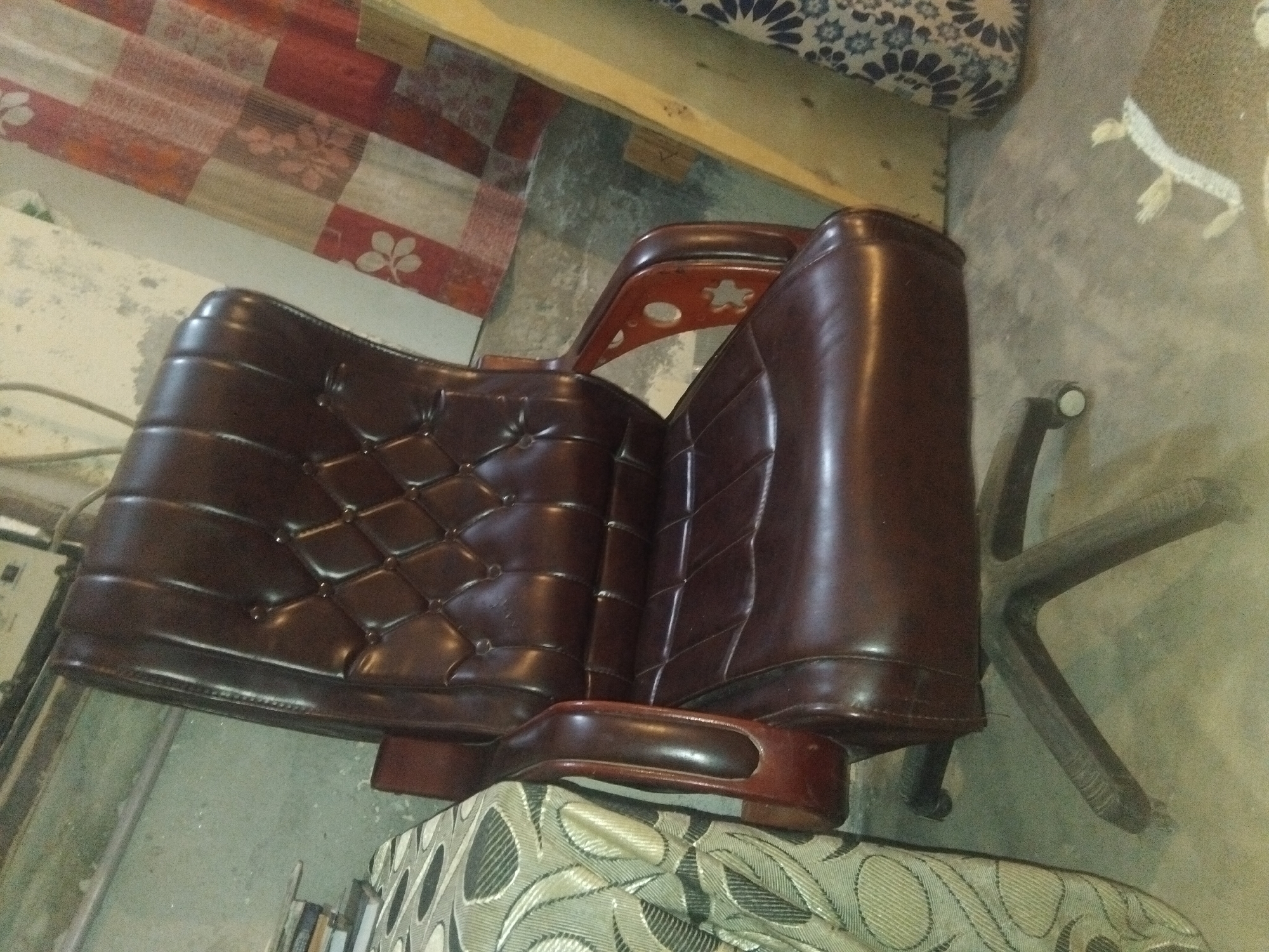 A brown leather executive chair with a high back and tufted cushioning sits on a five-wheeled base. The chair has wooden armrests with a curved design. To the left of the chair is a portion of a table covered with a patterned fabric, featuring an intricate leaf design in muted green and beige tones. In the background, a patterned curtain with blocks of red, white, and floral motifs hangs. The floor appears to be concrete, with some debris and a small area rug with a white floral design visible towards the bottom right. The overall environment suggests an indoor setting, possibly a home or office, in Haveli Jhangranwali, Pakistan.