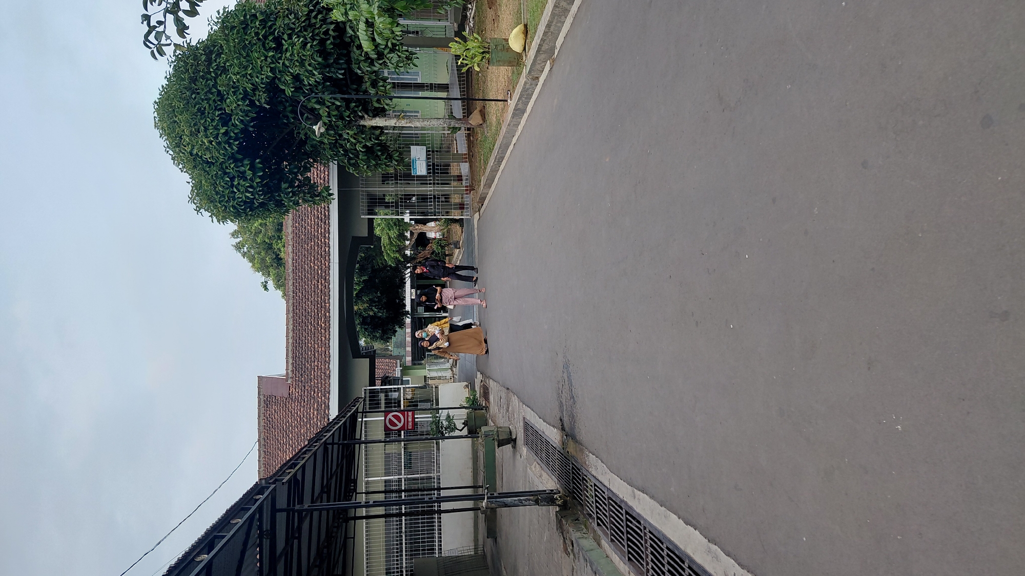 The image depicts an outdoor scene on a paved road within a complex, likely a medical facility or educational institution, in Rejasari, Indonesia. The road leads towards an arched gateway in the distance, flanked by buildings with tiled roofs and green facades.
In the foreground, on the left side of the road, is a covered walkway with a metal frame and a dark roof. Adjacent to this walkway is a drainage grate running along the edge of the road. A red and white sign with a diagonal line through a circle is visible on a pole near the walkway.
Further down the road, approaching the arched gateway, are four individuals. The leftmost individual is wearing a light brown, long dress and a headscarf. To their right, a person in a light yellow headscarf and a white top is walking. Next to them, a person in a light pink outfit, including a headscarf and pants, is walking. The rightmost individual is wearing a dark jacket and pants. They appear to be walking away from the viewer towards the gateway.
Beyond the gateway, more buildings and trees are visible. A large, dense green tree stands on the right side of the road, near the gateway, with a street lamp mounted on a pole nearby. The sky is overcast. The overall environment suggests a calm day in a well-maintained outdoor area.