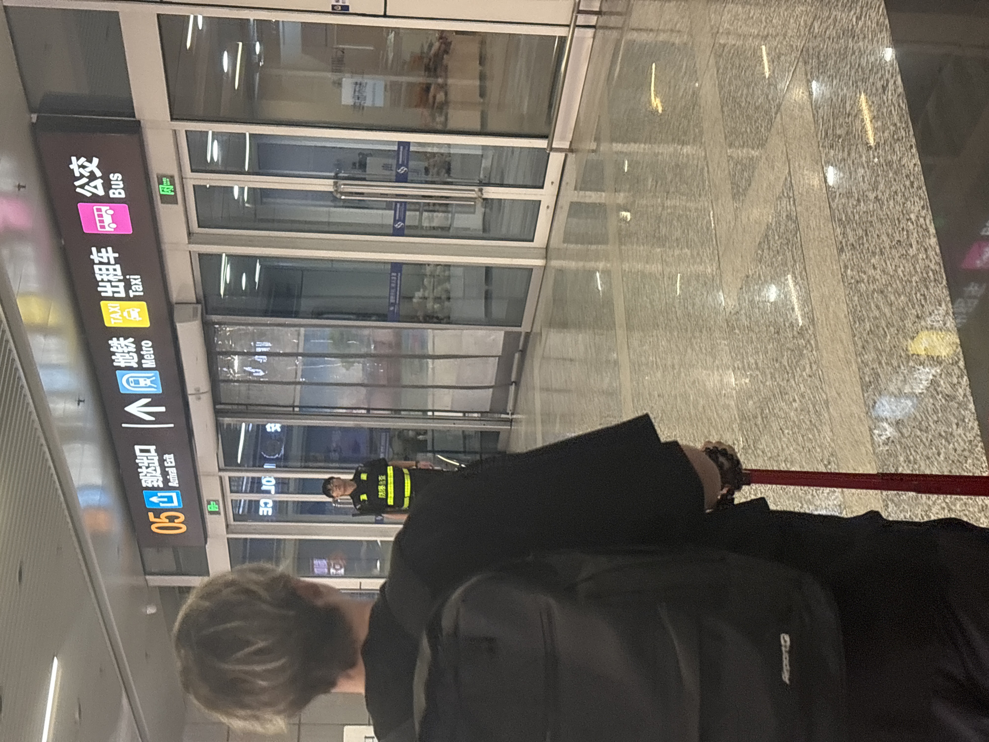 A person with blonde hair, wearing a black shirt and backpack, is seen from behind, holding a red suitcase handle. They are in an indoor environment, likely an airport or train station, with a polished, reflective floor. In the background, a sign displays "05 Arrival Exit," "Metro," "Taxi," and "Bus" in both Chinese and English, indicating transportation options. A security guard wearing a high-visibility vest is standing near the entrance. The location is identified as Xinwu District, China.
