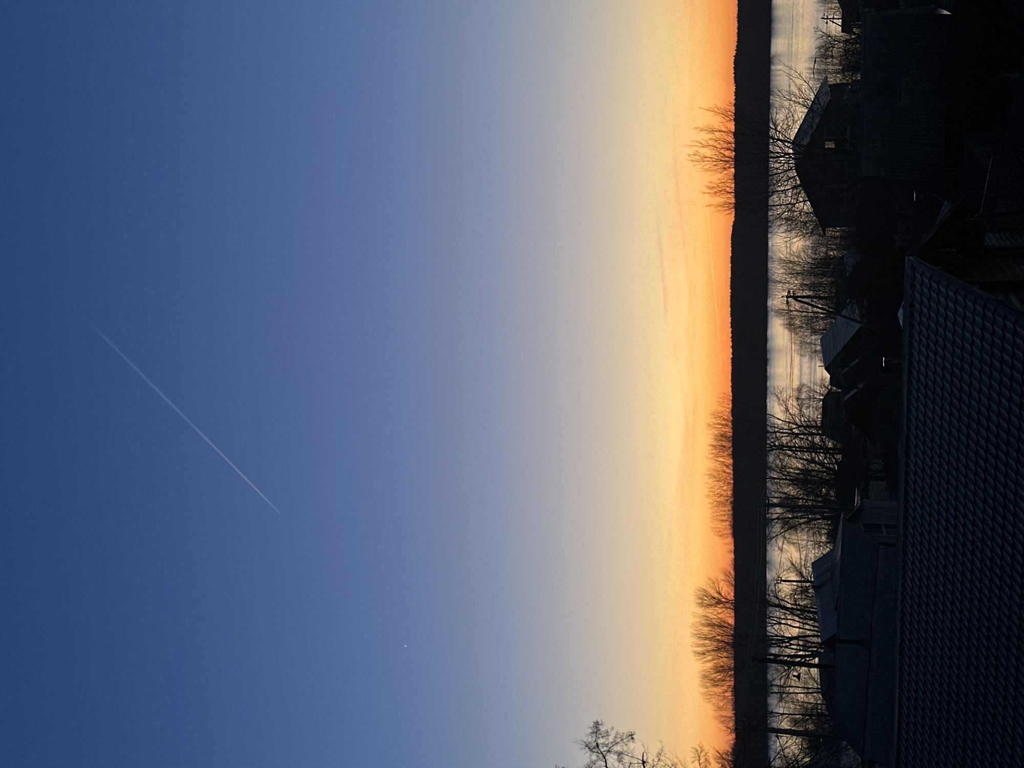 The image shows a sunset or sunrise over a body of water, possibly a lake or wide river. The sky transitions from a deep blue on the left to a gradient of orange and yellow near the horizon. A contrail is visible in the blue portion of the sky. Silhouetted trees line the horizon, and structures, including buildings with peaked roofs, are visible on the right side of the image. The image appears to be taken from an elevated position, perhaps from a building or high ground. The location is potentially Chaikovsky, Russia, based on the presence of houses.