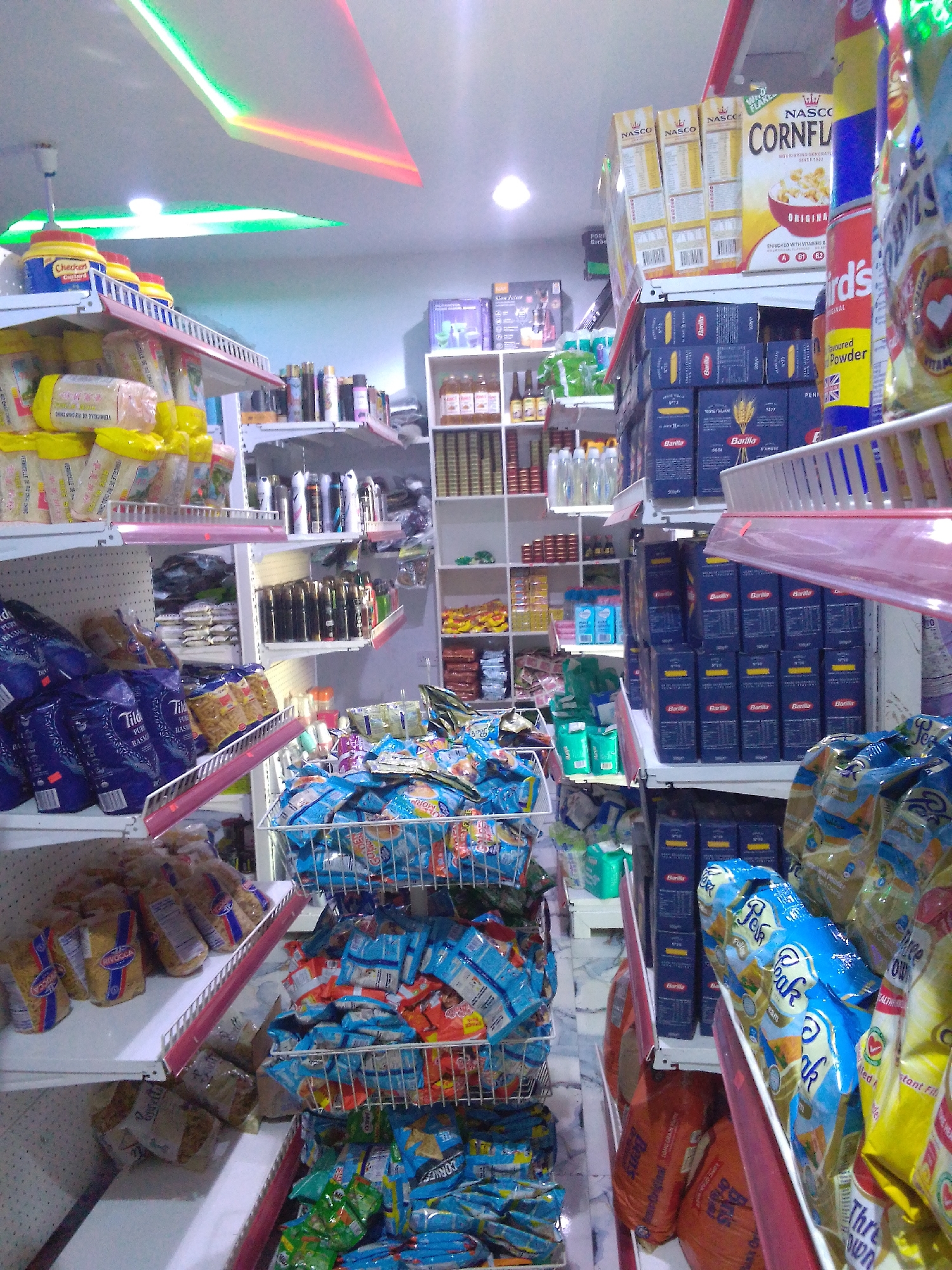 The image depicts the interior of a brightly lit convenience store in Howayi, Nigeria. The shelves are densely stocked with a variety of packaged goods, including pasta, cereal, and other food items. On the upper shelves, several boxes of "Nasco Cornflakes" are visible, along with various aerosol cans and bottled products. The lower shelves display large blue bags of rice, and a variety of bagged snacks and pastas in blue and yellow packaging. A metal shopping basket filled with blue-packaged snacks is also present. The store is illuminated by ceiling lights, including green and red strip lighting, and a fan is mounted on the ceiling. The overall impression is of a well-stocked retail environment. There are no people visible in the image.
