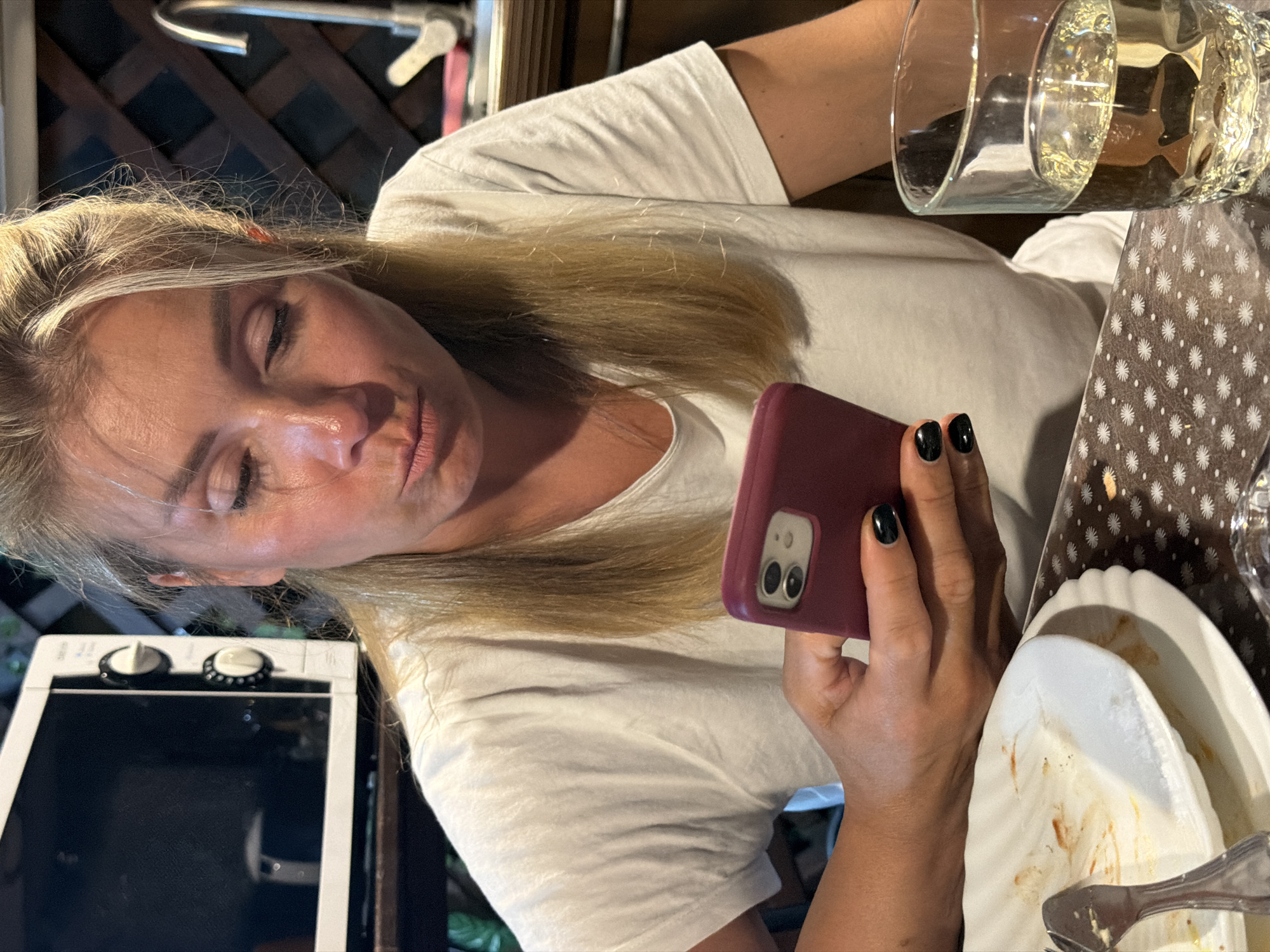 The image shows a woman with blonde hair, wearing a white t-shirt, looking at her phone. She is holding a maroon phone in her right hand, with black nail polish on her fingernails. In front of her, there are dirty plates with remnants of food and a glass of light-colored liquid, likely white wine. To the left of the woman, a white microwave is visible. In the background, a kitchen sink and faucet are present, along with a wooden lattice structure. The setting appears to be outdoors, possibly a veranda or patio, given the lighting and background elements, and the presence of a microwave suggests a casual dining or living area. The overall scene depicts a moment of leisure, with the woman engrossed in her phone while at a table with used dishes and a drink. The location of Sochi, Russia is not visually ascertainable from the image itself.