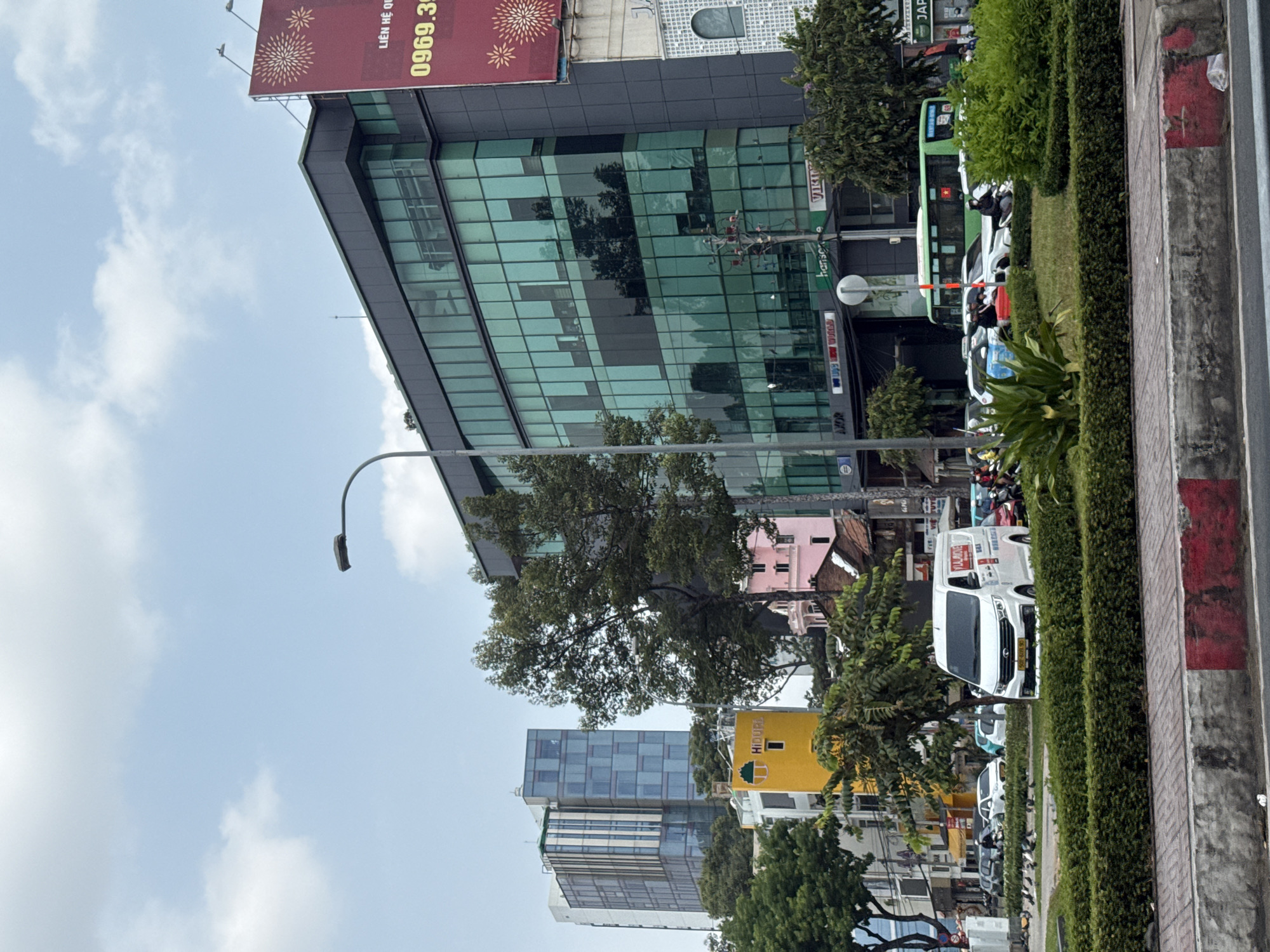 The image shows a street scene in Thủ Đức, Vietnam, with a mix of modern buildings and greenery. A tall glass-fronted building occupies a significant portion of the frame, with advertisements on its exterior. A variety of vehicles, including buses, vans, and motorbikes, are present on the road, suggesting traffic congestion. Sidewalks are lined with hedges and trees. The sky is visible with scattered clouds.