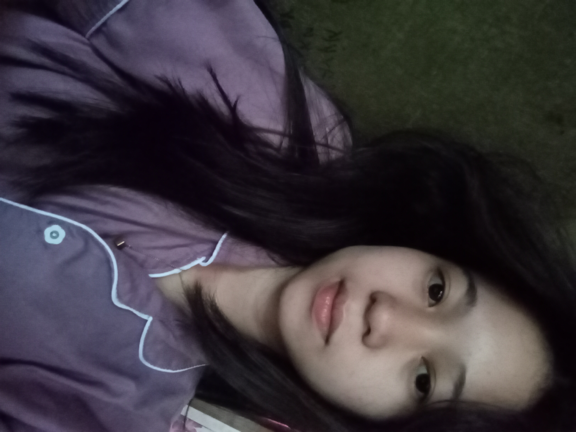 A young Indonesian woman with long, dark hair is pictured from the chest up, lying down and looking at the camera. She is wearing a light purple pajama top with white piping. The background is a blurred, dark green textured surface. The image appears to be taken indoors, possibly in a bedroom in Rejosari, Indonesia, given the subject's ethnicity and the potentially common domestic setting. There are no other individuals or objects of note in the frame. The lighting is soft and natural.