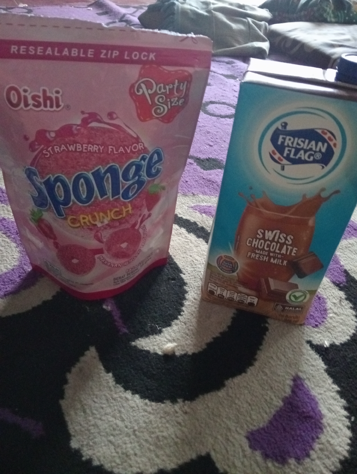 Two food products are placed on a patterned rug. On the left, a pink resealable zip-lock bag of "Oishi Sponge Strawberry Flavor Crunch" is positioned upright. The bag features the brand name "Oishi" in red lettering, "Strawberry Flavor" in script, and "Sponge" in large blue letters. A red graphic depicting a splash of liquid and strawberries is also visible. To the right, a carton of "Frisian Flag Swiss Chocolate" flavored milk is placed on its side. The carton has a blue and white logo with the words "FRISIAN FLAG" in a circle. Below the logo, an image shows a glass of chocolate milk being poured, with chocolate bars and the text "SWISS CHOCOLATE -MADE WITH- FRESH MILK". A green halal logo is also present on the carton. The background consists of a patterned rug with black, white, and purple accents. No people are visible in the image, and there is no indication of a specific location in Desa Silo, Indonesia.