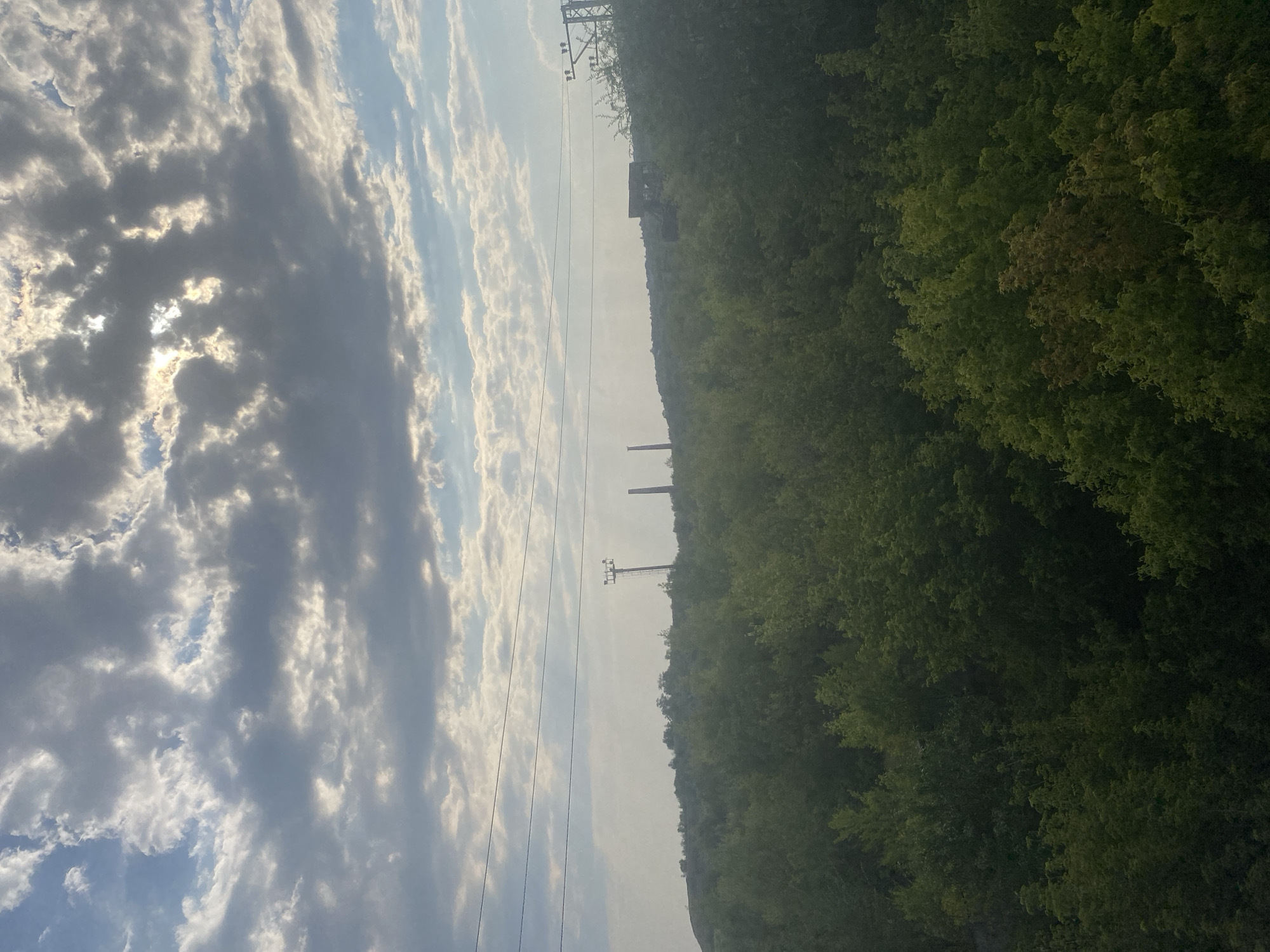 The image depicts a landscape dominated by a dense forest in the foreground and midground. In the distance, a hilly terrain is visible, topped with a few industrial structures and utility poles. The sky is filled with dramatic, cloudy formations, with sunlight breaking through in several areas, creating highlights and shadows. Several power lines traverse the scene horizontally. The overall impression is one of a rural or semi-industrial area, possibly on the outskirts of a city. The specific location in Oktyabrsk, Russia, is not visually evident from the image alone.