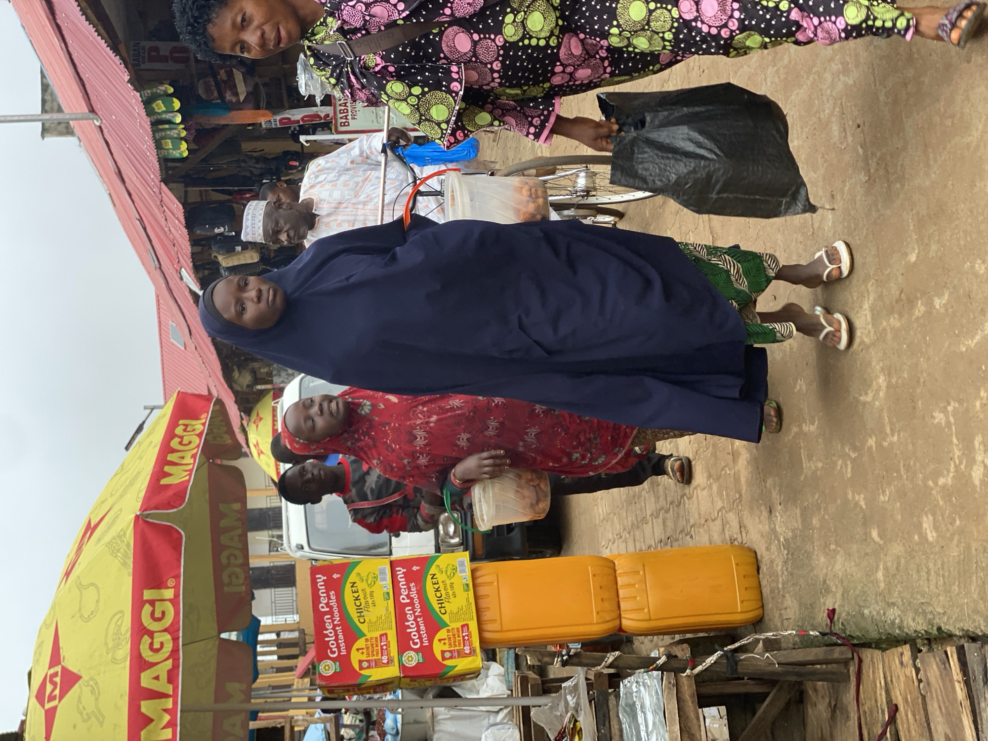 The image depicts a street market scene in Kafanchan B, Nigeria. In the foreground, a woman in a dark blue hijab walks towards the camera, carrying a large black plastic bag. To her right, another woman in a colorful patterned outfit walks in the same direction, also carrying a black bag. Behind them, a man wearing a white and orange striped garment, a white cap, and glasses is seen on a bicycle, holding a clear plastic bucket containing round, brown items. To the left of the frame, a young girl in a red headscarf and dress stands looking to the side, holding a similar bucket. In the background, a market stall displays stacks of yellow and red boxes of "Golden Penny Instant Noodles." An orange plastic drum is also visible. A yellow umbrella with the "MAGGI" logo shades part of the market. The ground is paved with rough, sandy-colored material, and buildings with red corrugated metal roofs line the street. The sky is overcast.