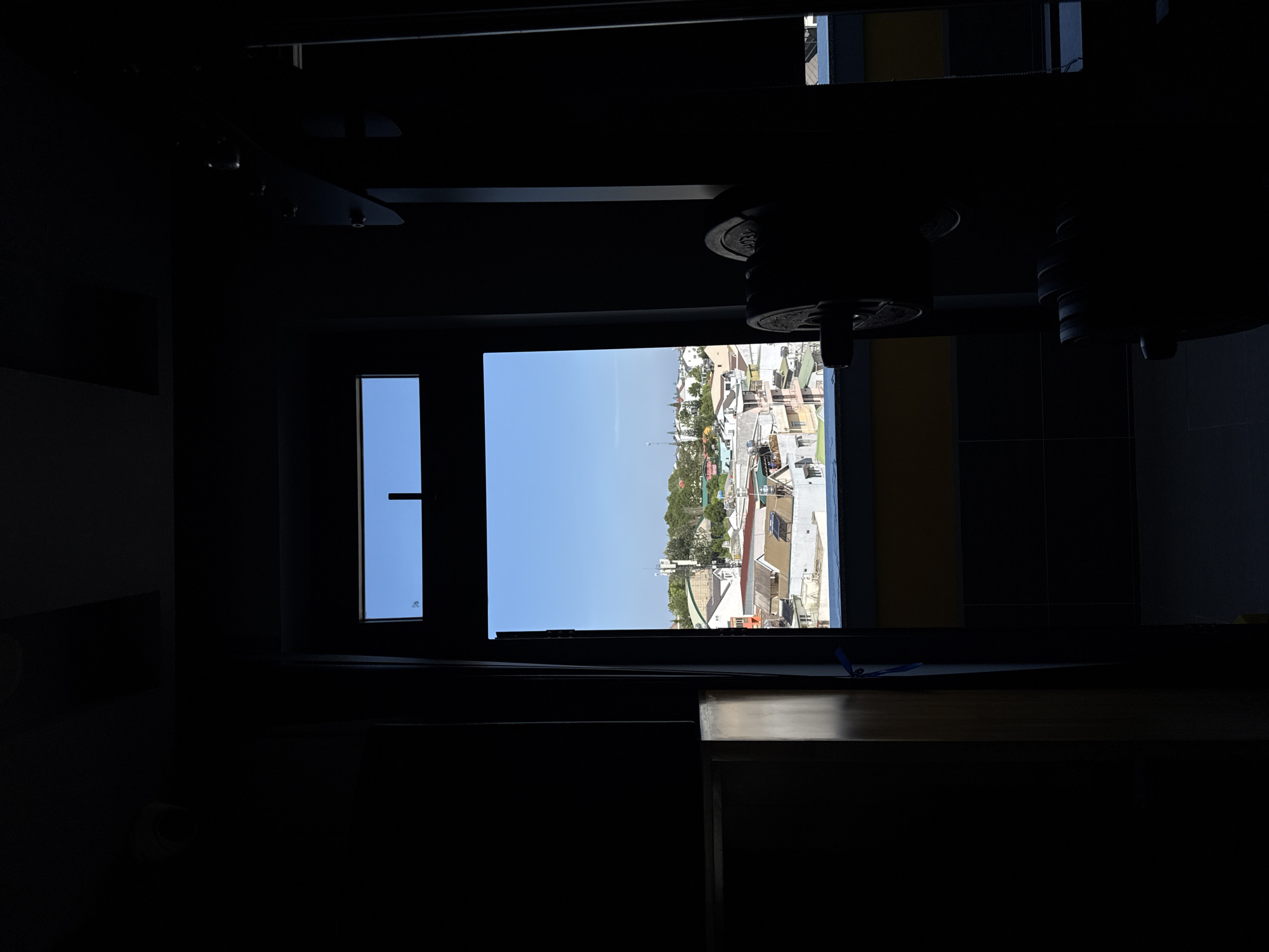 The image, oriented correctly, captures a view from a dimly lit indoor space looking out through a rectangular window. The room's interior is dominated by deep shadows, with dark ceiling structures and what appear to be fixtures vaguely discernible. Below the window, a surface, possibly a counter or table, is mostly in shadow, save for a narrow beam of sunlight highlighting a blue ribbon or cord.
Beyond the window, a clear, bright blue sky stretches over a bustling townscape, indicating it is daytime with sunny weather. The view encompasses a dense arrangement of buildings featuring various roof types, such as terracotta tiles and corrugated metal, many equipped with water tanks. Green foliage from trees and bushes is interspersed throughout the urban environment, suggesting a lush setting. In the distance, a tall, slender structure, likely an antenna or cell tower, stands prominently against the sky, with undulating hills or mountains visible further back. This architectural style and natural landscape are consistent with the provided location context of Lâm Viên - Đà Lạt, Vietnam.
No specific activity or event is taking place; the image presents a static, observational scene. The stark contrast between the bright exterior and the shadowy interior emphasizes the light source from the window. Faint, unreadable text or a logo is present on the lower left pane of the window glass.