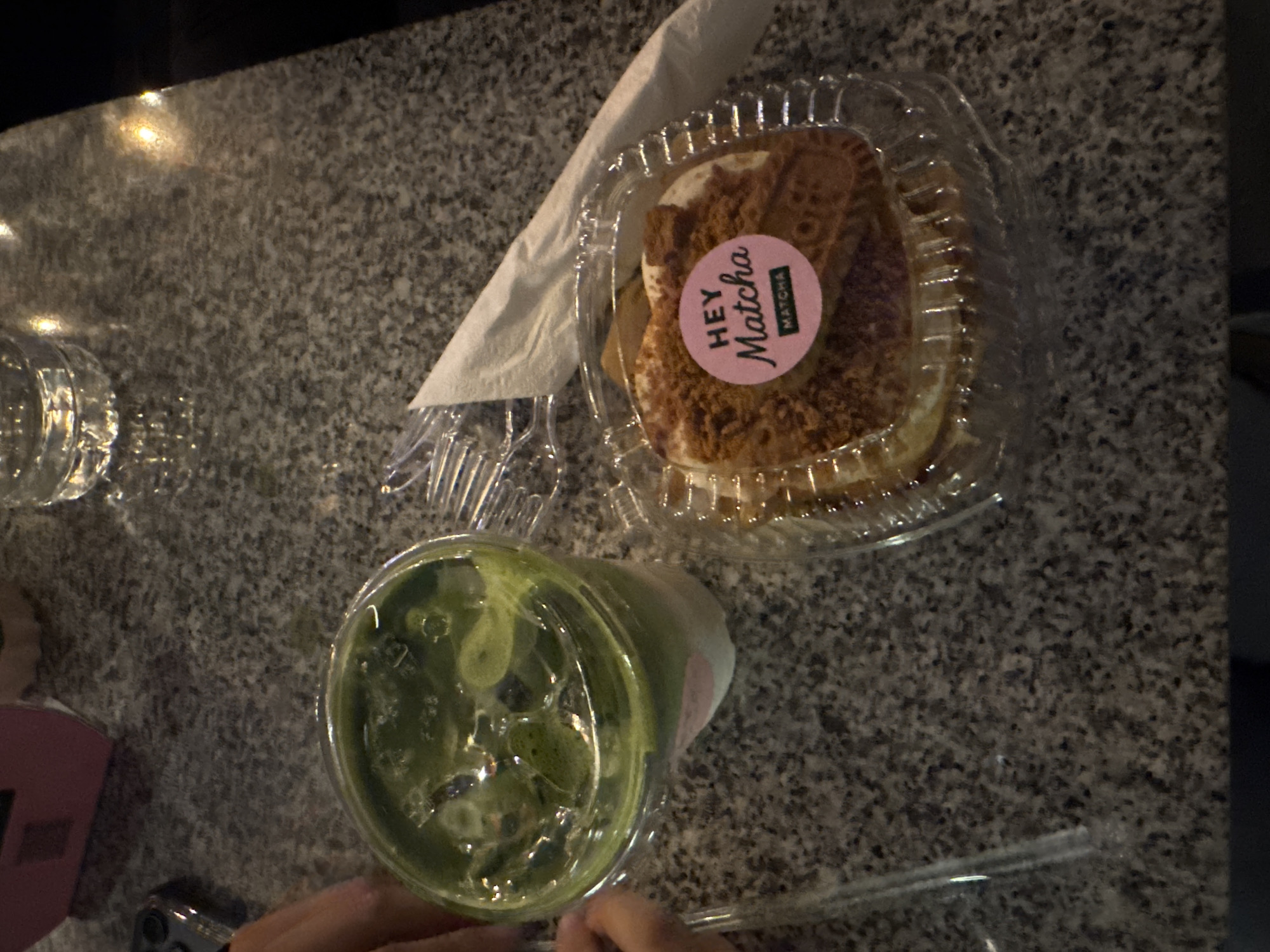 The image shows a gray speckled surface, possibly a countertop, with a partially visible water glass on the left. A folded white napkin lies between the glass and a plastic container holding a pastry topped with crumbs and a pink "Hey Matcha" sticker. Below the container, a clear plastic cup filled with a green liquid, likely a matcha-based drink with ice, is visible. A clear straw is present near the bottom of the cup. A person's hand is partially visible near the bottom left corner.
The image was captured in San Bernardino Tlaxcalancingo, Mexico.