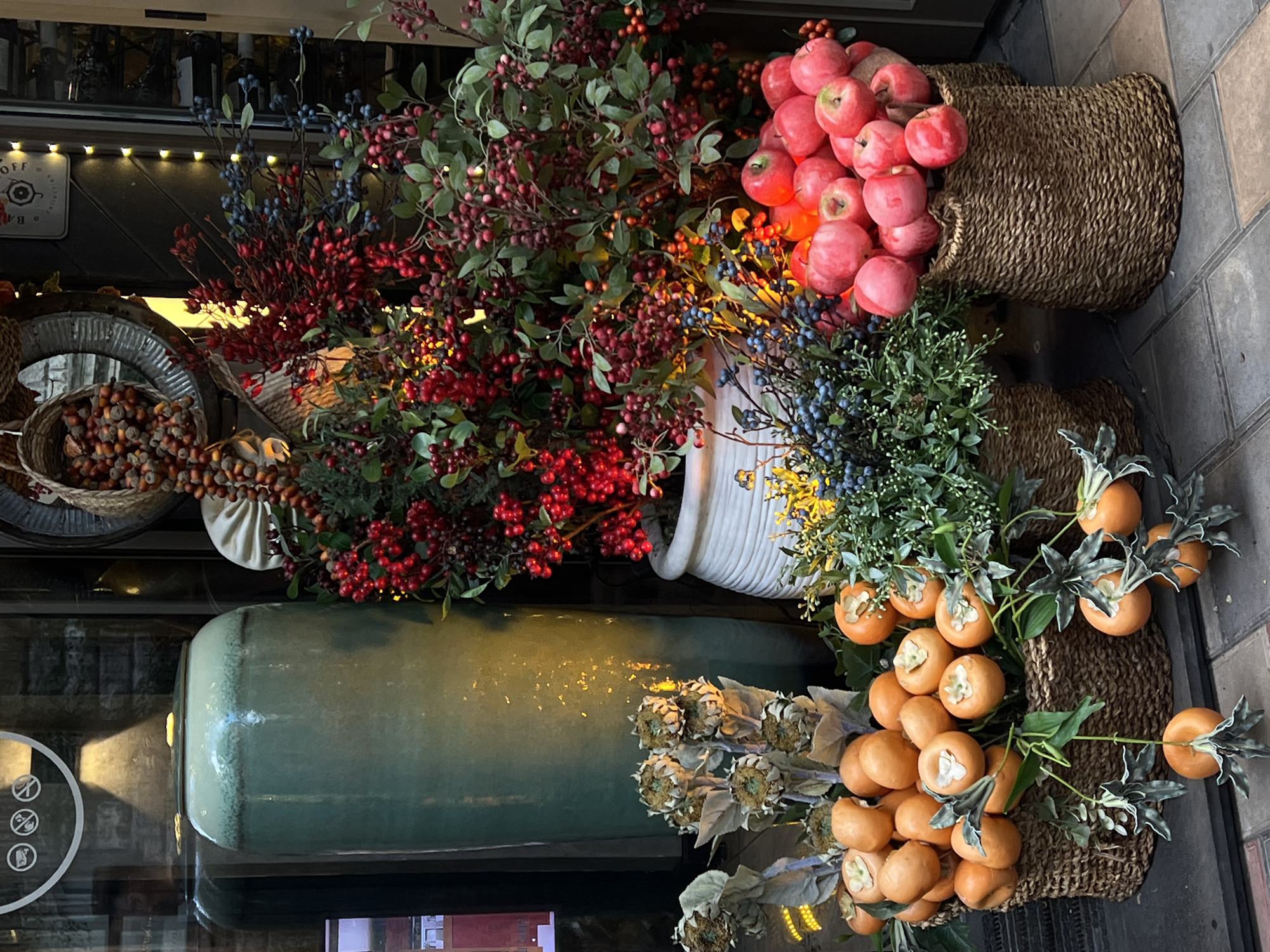 This image captures a vibrant, autumnal or winter-themed decorative display, likely at the entrance or window of a commercial establishment in Yerevan, Armenia. The scene is filled with a profusion of artificial fruits and berries arranged in various containers.
In the upper right, a large woven basket overflows with bright red, faux apples, illuminated by warm yellow string lights that cast a soft glow. Below this, another basket holds blue and green berry branches. A third, prominent woven basket in the foreground is filled with numerous orange, persimmon-like fruits, accompanied by green foliage and dried, sunflower-like decorative elements.
To the left, a tall, glossy teal-green ceramic vase stands, reflecting light. Behind it, a white ribbed vase holds an assortment of red and blue faux berry branches. Further left, a distressed metal container with a slightly reflective surface is filled with brown acorns and small, round decorative objects, accompanied by greenery.
The background reveals a dark interior, possibly a shop, with bottles visible on shelves and warm string lights creating an inviting ambiance. The ground beneath the display is paved with light grey blocks. Notable textual details include partial words "OFF" and "BAL" on a sign in the background. A round decal on a glass surface displays three crossed-out pictograms: one for no smoking, one for no food/drinks, and another likely for no pets or large bags, indicating entry restrictions. The overall impression is a festive and richly textured seasonal display.