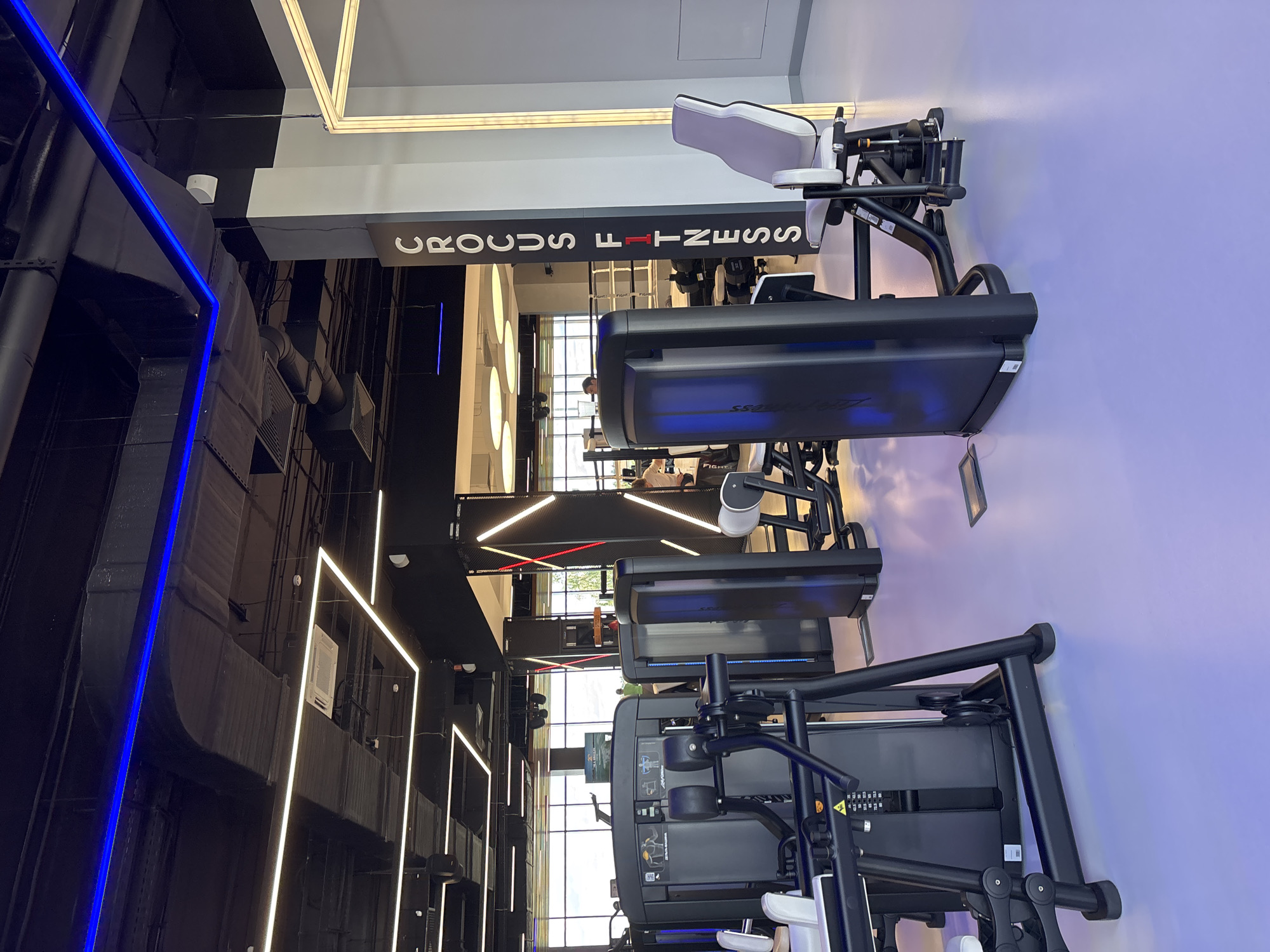 The image depicts the interior of a modern gym, identified as "CROCUS FITNESS" by signage on a pillar. The gym features several pieces of weight training equipment, with a focus on the front-left area where a black weight machine with pulleys is prominently displayed. In the background, more weight machines and workout stations are visible. The environment is characterized by a sleek design with black ceilings featuring industrial ductwork and a combination of blue and white LED lighting strips. The floor is a smooth, light-colored surface with a subtle purple hue, likely due to the lighting. The overall impression is that of a clean, well-equipped, and contemporary fitness facility. While there are some indistinct figures of people in the background, no specific actions or interactions are clearly discernible. The location within the city of Monolit, Russia, is provided as context.