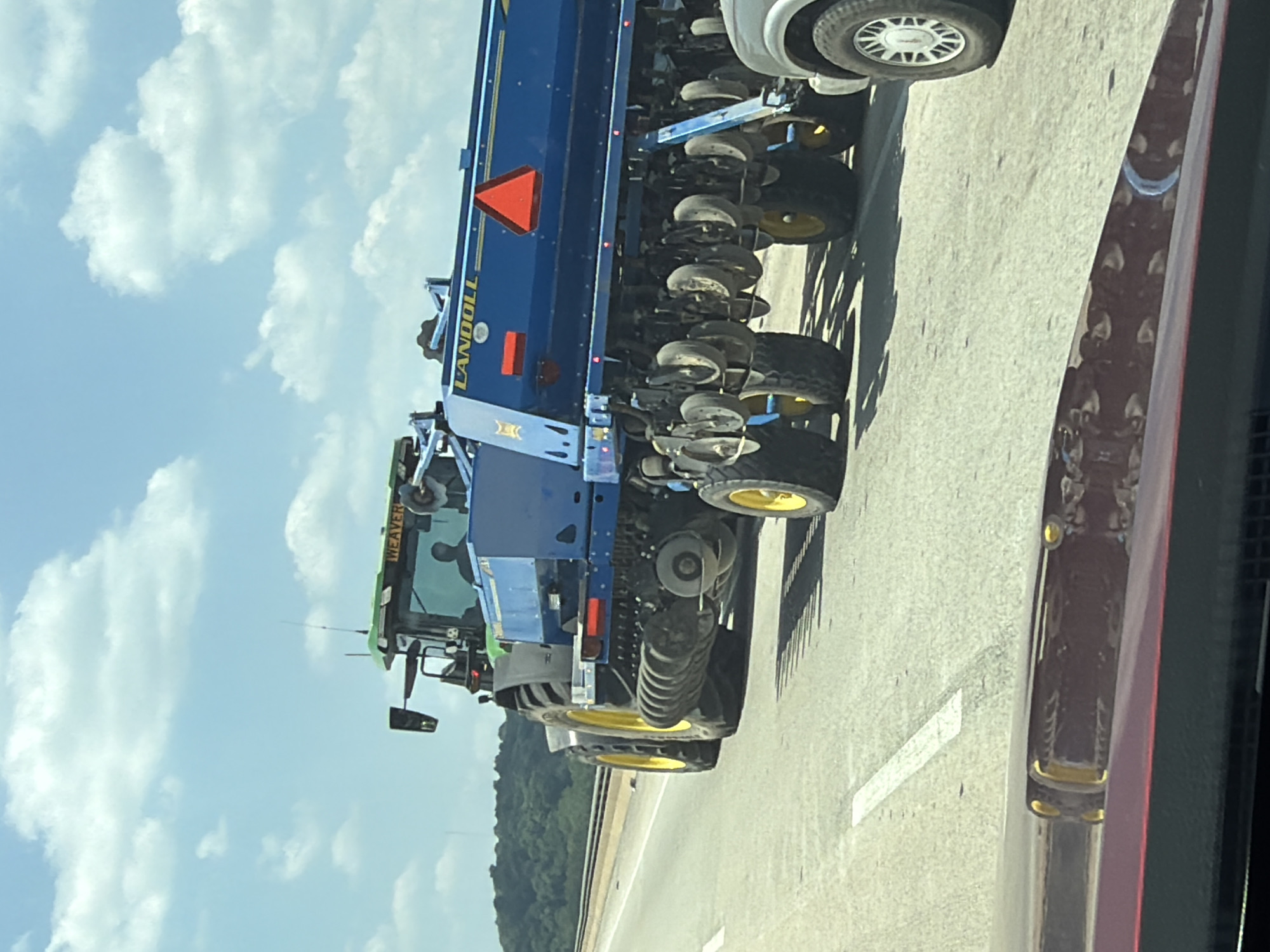A green and black tractor, possibly a John Deere, pulls a large blue agricultural implement with the brand name "LANDOLL" visible in yellow lettering. The implement is equipped with multiple seeding or planting units, each with small wheels. The tractor and implement are traveling on a paved highway, indicated by lane markings. In the background, a dense line of trees forms the horizon, against a sky with scattered white clouds. The scene appears to be in a rural or semi-rural area, given the agricultural machinery and natural landscape.