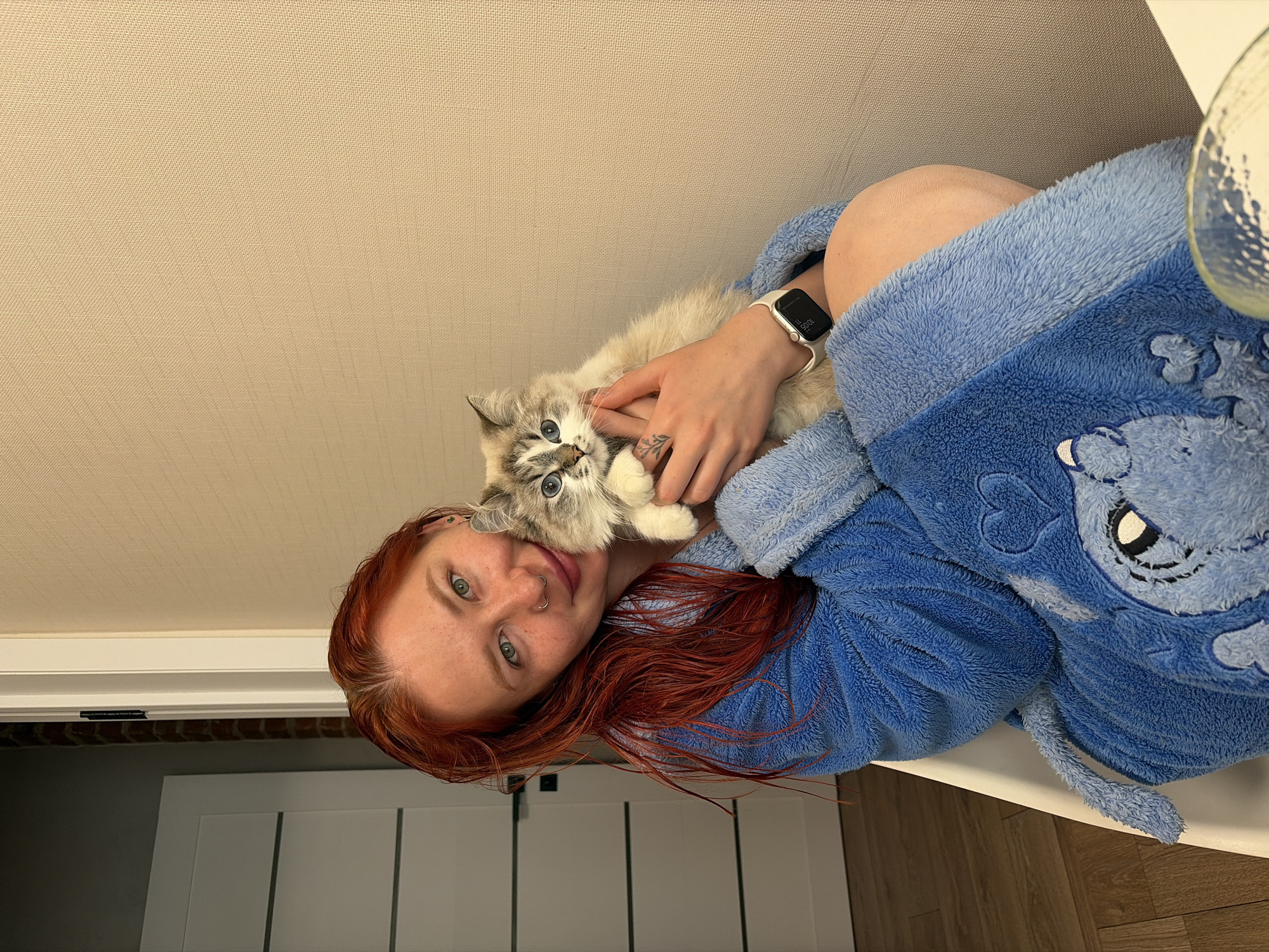 A fair-skinned woman with red hair is holding a kitten in her arms. She is wearing a blue robe. A smartwatch is visible on her wrist. The location is indoors with a white door and a light-colored wall. The exact location in Rostov-on-Don, Russia, cannot be determined from the image.