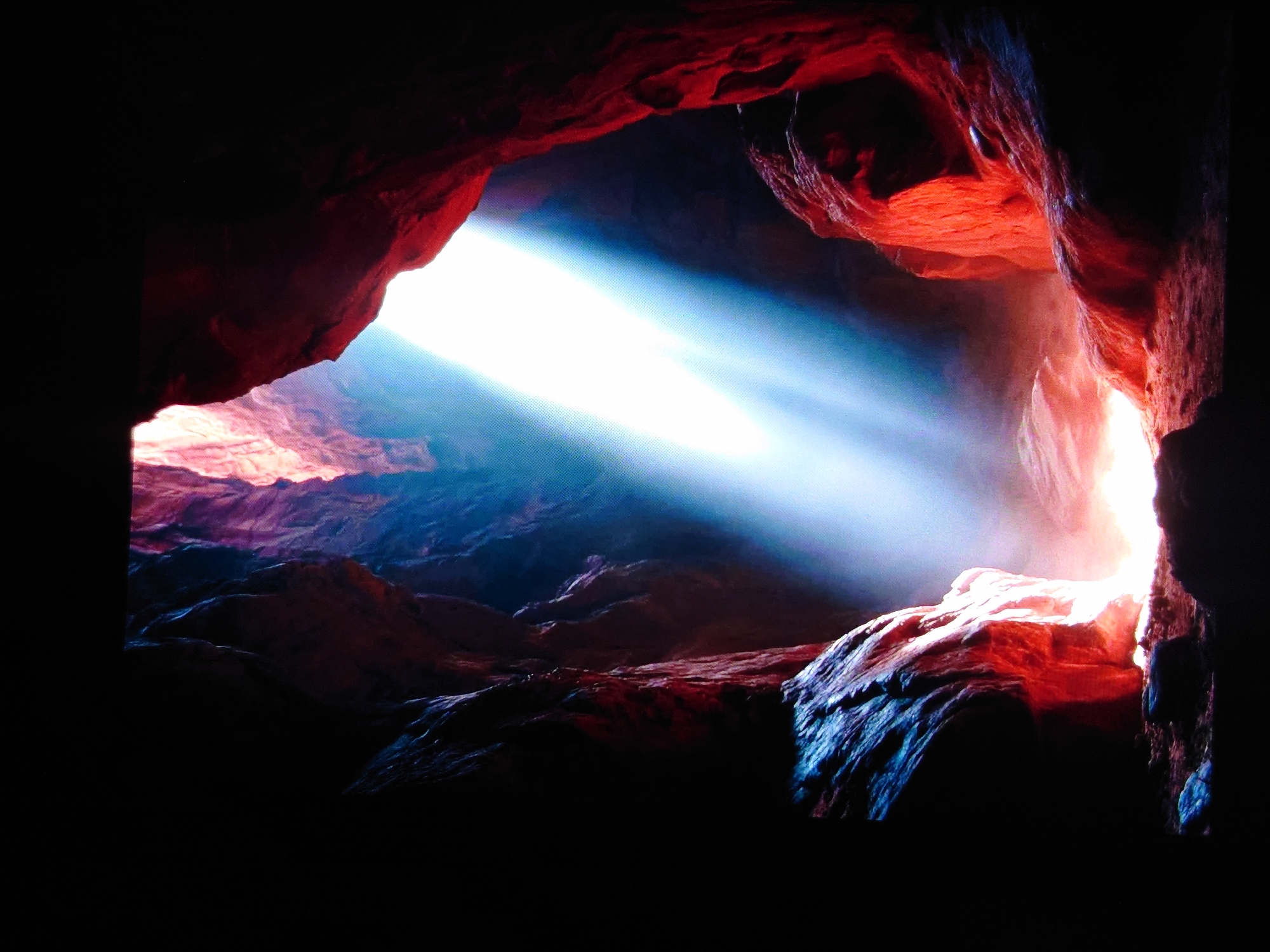 The image depicts a narrow canyon or cave with sheer rock walls. A strong beam of bright, white light, possibly sunlight, enters from the top of the opening and illuminates a portion of the canyon floor, creating a hazy effect with dust or mist. The rocks are predominantly in shades of red and orange, with some areas appearing darker or tinged with blue in the shadows. The overall impression is one of natural geological formation, dramatic lighting, and a sense of depth within the enclosed space. There are no people, animals, or man-made objects visible in the image. The scene is a natural landscape. There is no information within the image or accompanying text to suggest a specific location in Belorechensk, Russia, or any other city.