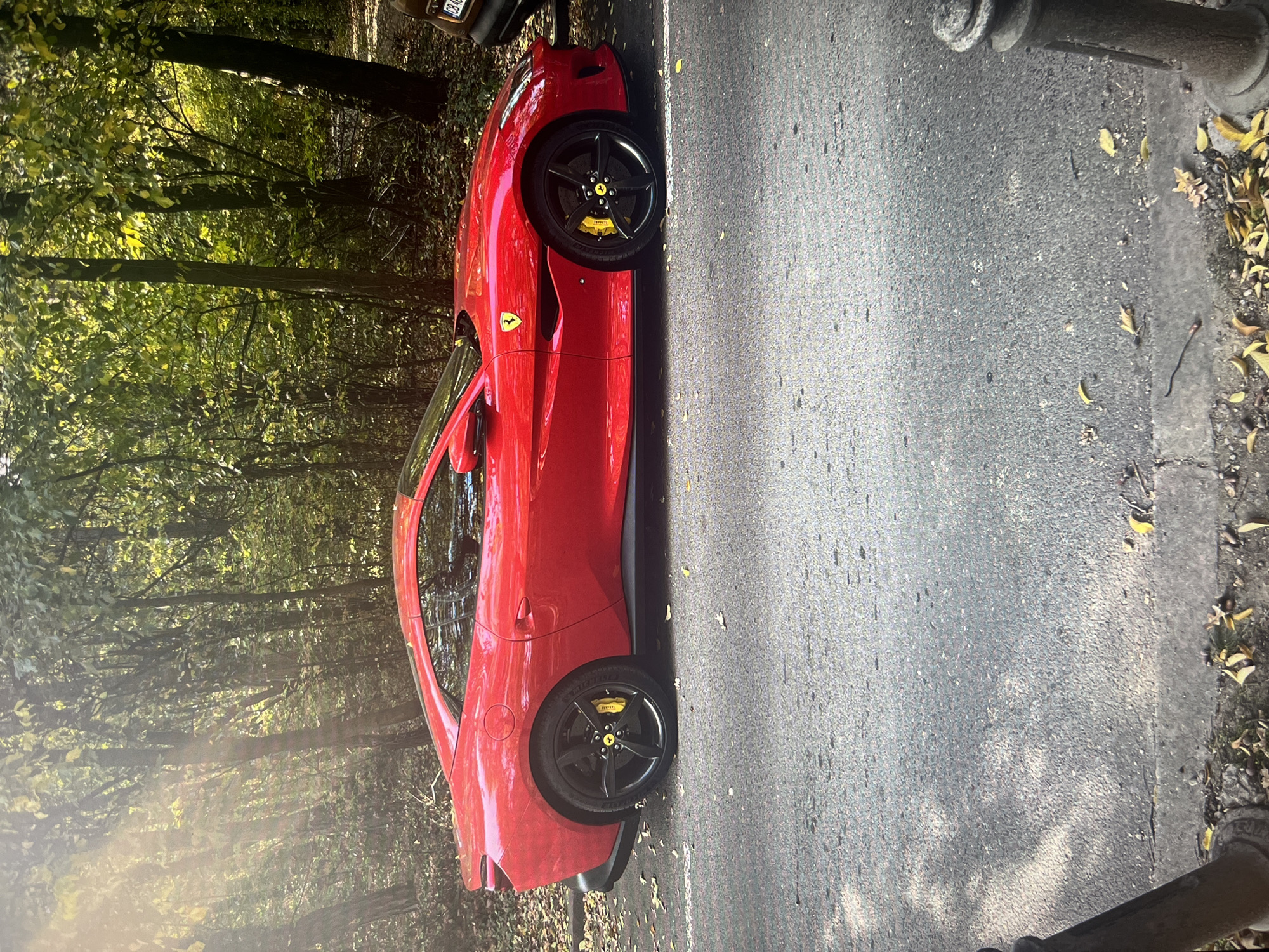 A red Ferrari is parked on the side of a road near a wooded area, likely in a park or residential area. The car has black wheels and yellow brake calipers. The road surface is asphalt and there are trees and foliage in the background. Leaves are scattered on the ground along the roadside.
