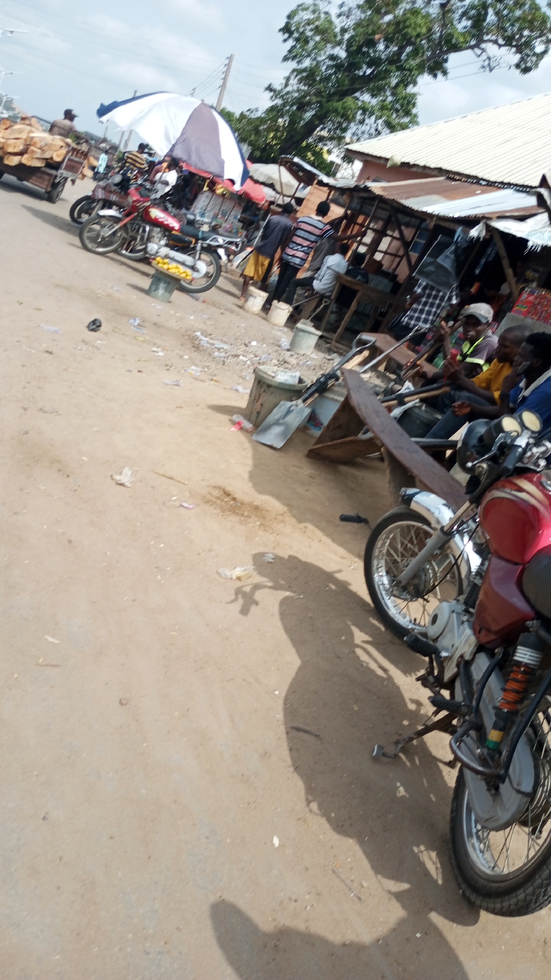 The image depicts an outdoor scene at Zango-ndaji, Nigeria. In the foreground, there's a red motorcycle parked on a dirt road. The midground shows a group of people gathered near a row of market stalls with corrugated metal roofs. Some individuals are seated, while others are standing and appearing to conduct business. There are also several motorcycles parked nearby, some with umbrellas attached. In the distance, a cart loaded with logs is visible. Debris is scattered on the ground.