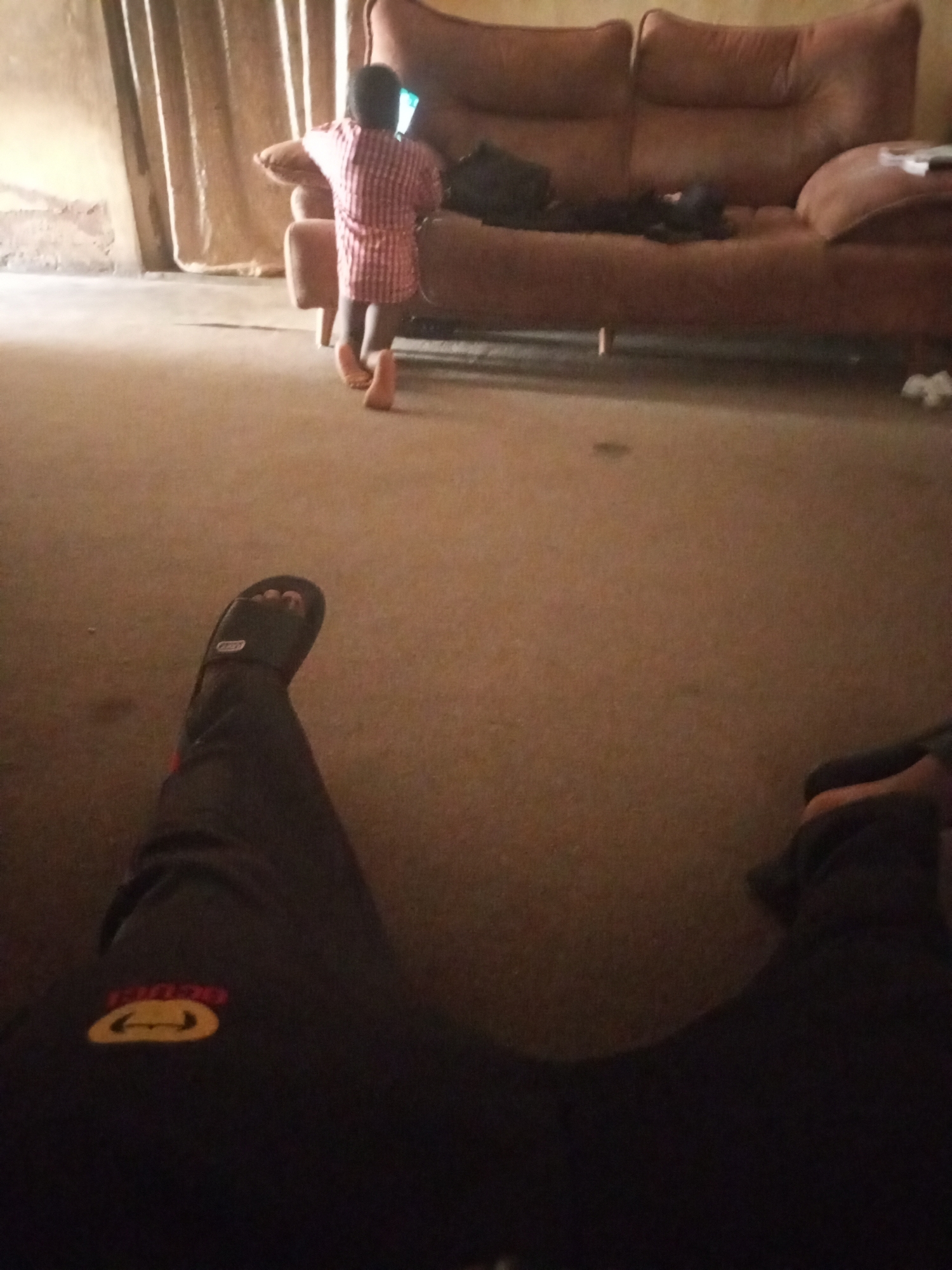 A young child wearing a red and white striped shirt is kneeling on the floor in front of a brown couch, looking at a phone. The child's back is to the camera. The couch has several pillows and is positioned against a wall with a beige curtain. The floor appears to be carpeted in a brown hue. The foreground of the image is dominated by a pair of legs wearing black pants and black slides, suggesting the photo was taken from a seated or reclining position. The environment appears to be indoors.