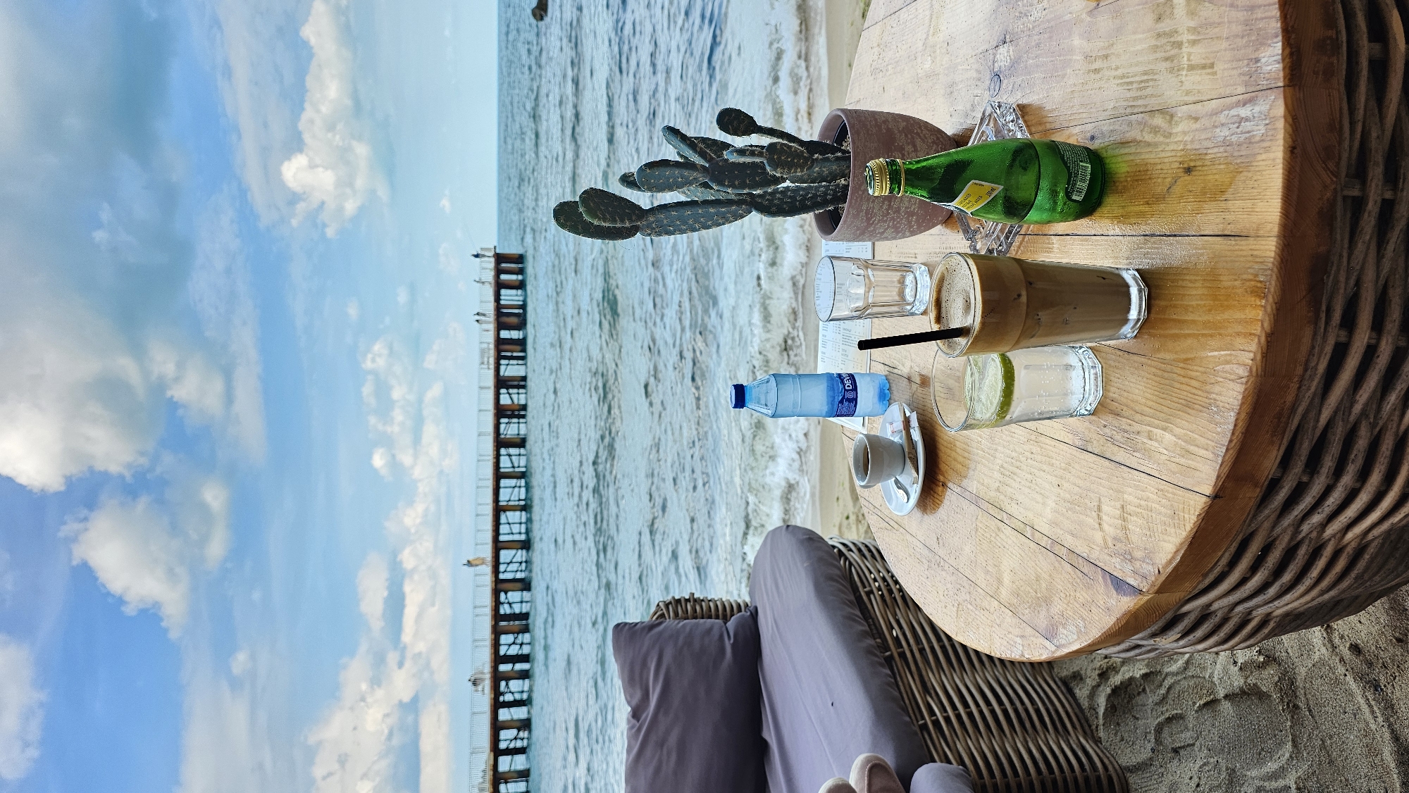 The image depicts a table setting at a beachside location, likely a cafe or restaurant. The table is round and wooden, with a textured edge resembling wicker. The setting includes a cactus in a pot, a glass bottle of Perrier water, an empty glass, a glass of iced coffee with a straw, a glass of a lime cocktail, a bottle of water, and a cup of coffee on a saucer. The table sits next to a cushioned chair. In the background is a view of the sea with a long pier stretching into the water. The sky is visible with clouds. Based on the pier style and typical coastal scenery, this image likely captured in Varna, Bulgaria. No people are in view, apart from a pair of feet at the bottom of the image.