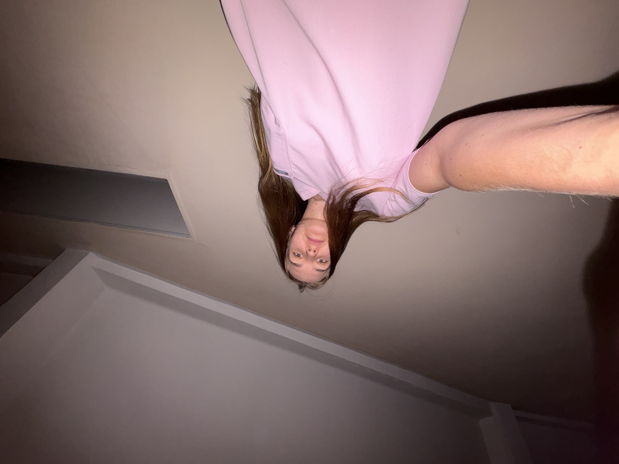 The image displays a young person, presumably female, captured in an unusual, upside-down orientation within an indoor setting. The individual has long, dark brown hair flowing downwards and is wearing a light pink short-sleeved top. Her head is positioned at the top center of the frame, with her face looking directly at the camera with a neutral expression.
The scene appears to be an interior room, likely residential or commercial, characterized by plain, light-colored walls and a ceiling. The perspective suggests the camera is positioned below the person, looking upwards, or the person is lying down and holding the camera above themselves, resulting in the inverted view. In the background, there's a rectangular recessed area or opening on the left side, possibly an air vent or a design feature. Trim or molding is visible along the upper edges of the walls where they meet the ceiling.
The lighting is dim and artificial, indicating an indoor shot, possibly during the evening or night, or in a poorly lit room. No direct sunlight or outdoor elements are visible. There is no legible text on the person's clothing or in the background, though a small, unidentifiable tag is present on the pink top. No specific visual cues within the image directly relate to the Perm, Russia location context.