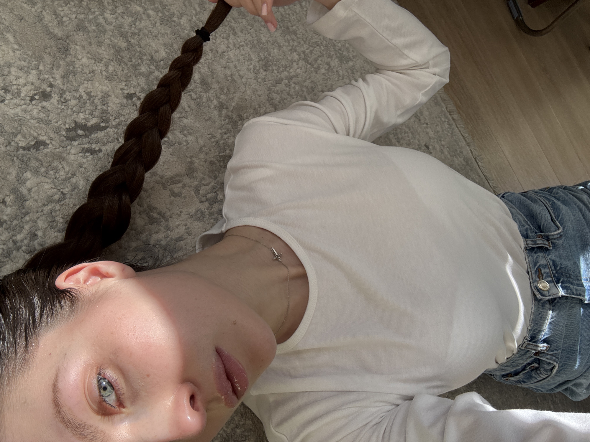 A young woman is lying on a grey, textured carpet. She is wearing a white, long-sleeved shirt and light blue jeans. Her hair is dark brown and braided, held together with a black elastic band. She is wearing a delicate silver necklace with a small pendant. The location is indeterminate and there is no clear indication that it is Moscow, Russia. The light source appears to be natural, with a patch of sunlight falling on the floor and her face.