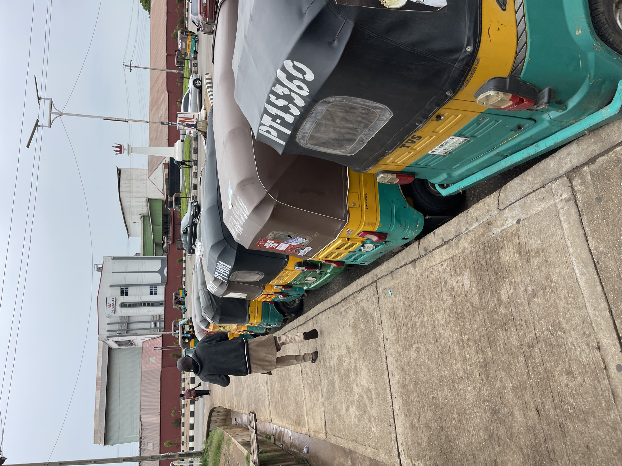 The image shows a row of parked tricycles, known locally as "Keke Napep", along a roadside in Jos, Nigeria. The tricycles are predominantly yellow and green, with registration numbers visible on the covers. A man is walking along the sidewalk beside the tricycles, wearing a dark jacket and tan pants. In the background, there is a building with the name "NASCO" on it, suggesting the location is near the NASCO factory or headquarters in Jos. Utility poles and wires are visible in the sky.