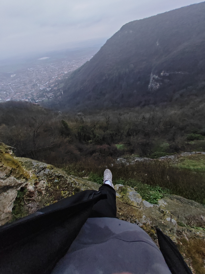 The image presents an overhead perspective from a rocky outcrop overlooking a valley containing the city of Shumen, Bulgaria, under an overcast sky. In the foreground, the lower half of a person is visible, wearing a dark grey cap or hood, a dark jacket, and black pants. Their right foot, clad in a light grey sneaker with white laces, rests on the moss-covered rock ledge at the edge of the cliff. Below and extending into the midground are steep, vegetated slopes, primarily covered with leafless trees and shrubs, descending into a deep valley. The city of Shumen, characterized by dense residential and commercial buildings, is visible in the distance across the valley. A large, dark, forested mountain face with visible rocky sections occupies the right side of the background, further framing the valley. The sky above is uniformly grey and cloudy.