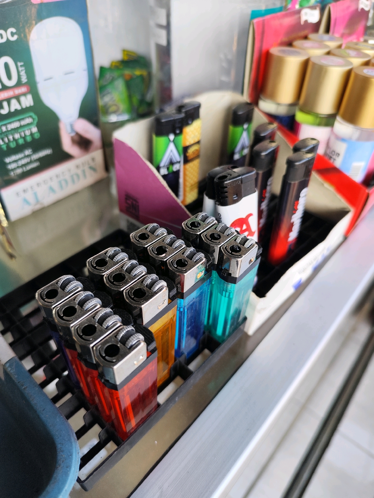 A close-up, eye-level shot captures a display of lighters and other items in what appears to be a small shop or market stall. In the foreground, a row of transparent lighters in various colors, including red, orange, green, and blue, are neatly arranged in a black plastic holder. These lighters have silver metal tops with a rotating wheel for ignition.
Behind the lighters, a mix of products is visible. To the left, a cardboard box advertises an "EMERGENCY BULB ALADDIN" with an image of a large LED bulb. Further back, in a pink and white display, are several black and green lighters, some with distinctive logos. In the background, shelves hold more items, including what appear to be small bottles with gold and silver caps, possibly cosmetics or fragrances, and some green packaged goods.
The overall environment suggests a retail setting with a variety of small consumer goods. The lighting is bright, highlighting the colors and details of the products. There are no people clearly visible in the shot, and the focus is on the arrangement and presentation of the merchandise. The exact location in Bambanglipuro, Indonesia, is not visually indicated within the image itself.