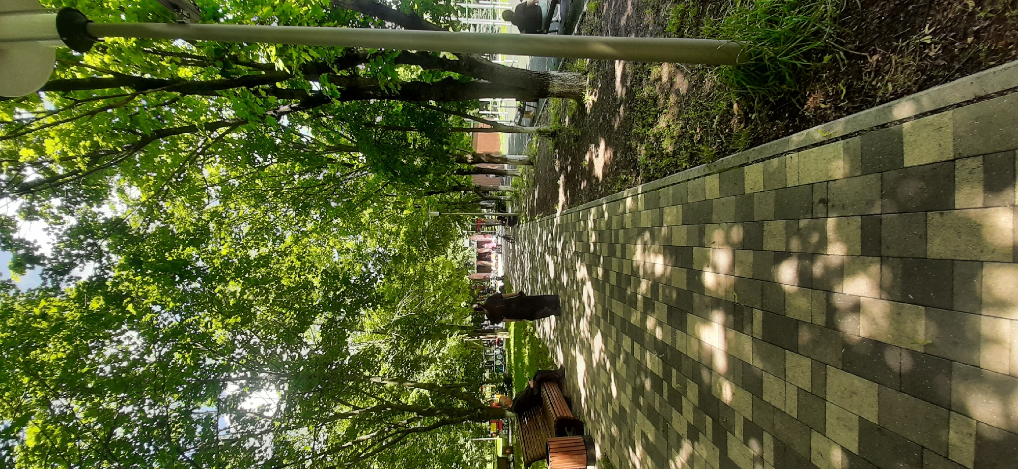 The image depicts a pathway in a park-like setting, possibly in Yessentukskaya, Russia. The pathway is paved with square tiles of alternating dark grey and light beige colors, with dappled sunlight filtering through the overhead tree canopy, casting shadows on the ground. To the left of the pathway, a wooden bench is occupied by a person sitting with their back to the camera, and further down the path, a lone figure in dark clothing walks away from the viewer. The right side of the pathway features a grassy area with young trees, and a lamppost is visible. In the background, there are structures that appear to be part of a park's amenities, including what looks like a small train and playground equipment, with other park visitors in the distance. The overall atmosphere is one of a pleasant, sunny day in a public recreational space.