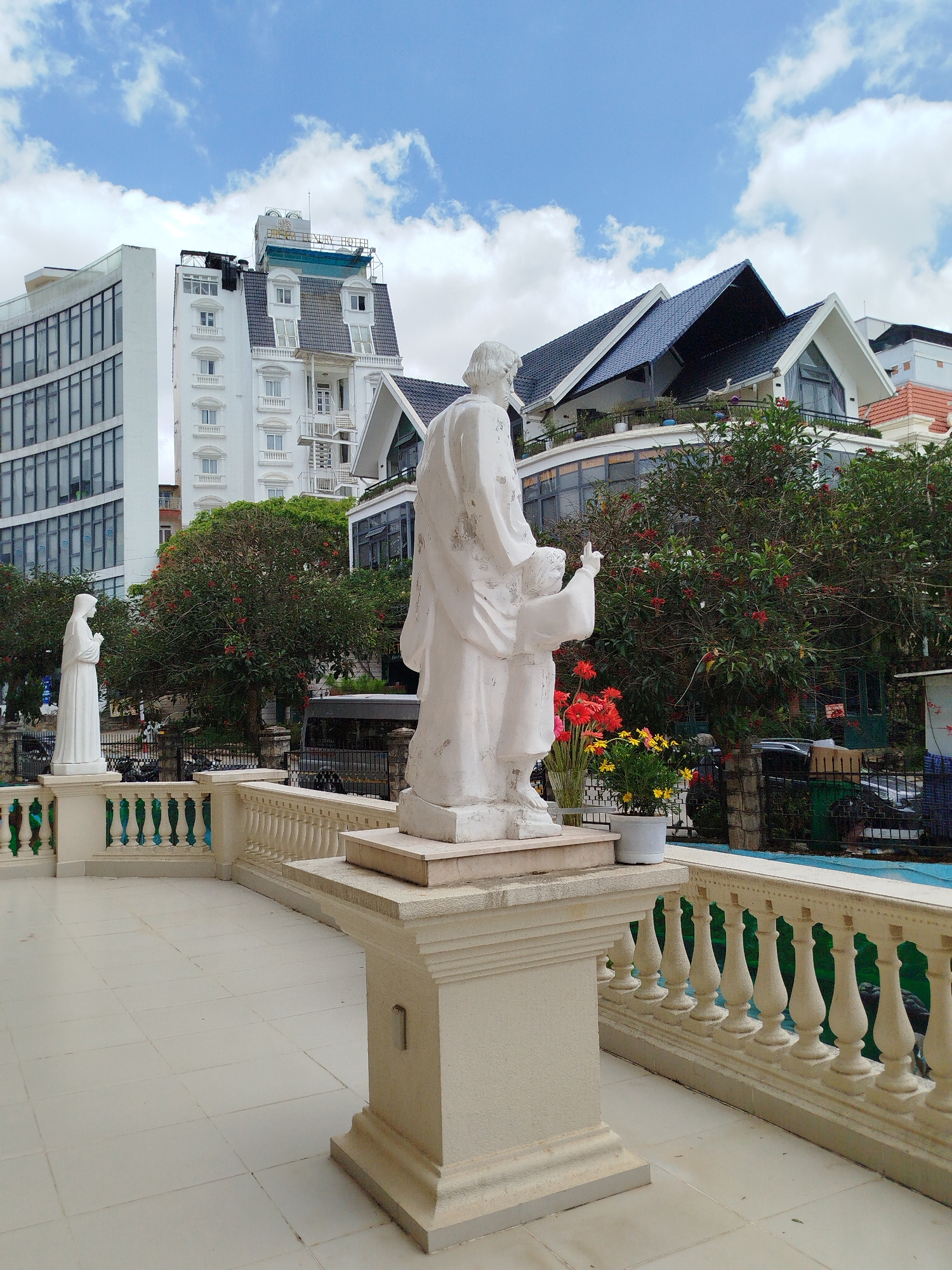 The image shows two white statues, with a larger statue of a man and child standing on a platform in the foreground and a smaller statue of a woman in the background, against the backdrop of modern buildings and a partly cloudy sky in Da Lat, Vietnam.
The statue in the foreground depicts a man with a child by his side, both rendered in white. The man has his arm raised, while the child looks up at him. The statue is placed on a beige platform with a rectangular base. To the side, are potted plants with red and yellow flowers.
In the background, modern buildings of various architectural styles can be seen, including structures with curved glass facades and high-pitched roofs. Trees with red flowers are interspersed between the buildings. A white balustrade with green accents lines the edge of the tiled terrace where the statues are positioned. A secondary statue of a standing woman in a white robe is present on a raised pedestal behind the main statue. The scene takes place in daylight, with the sky filled with blue patches and white clouds.