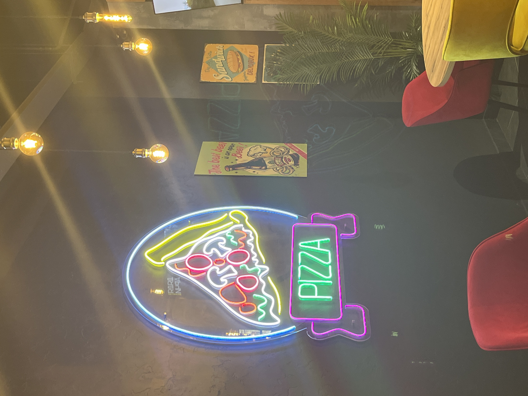 The image shows the interior of a restaurant, possibly a pizzeria, featuring a neon pizza sign with the word "PIZZA" in vertical green letters. The sign is mounted on a dark wall adorned with vintage-style advertisements. A red chair is visible on the right side of the frame, near a round wooden table. There are several hanging light bulbs at the top. The restaurant is located in Izhevsk, Russia, as evidenced by the inscription "RIRI INPGA" on the pizza sign. The environment is dimly lit, with the neon sign and light bulbs providing the main sources of illumination.
