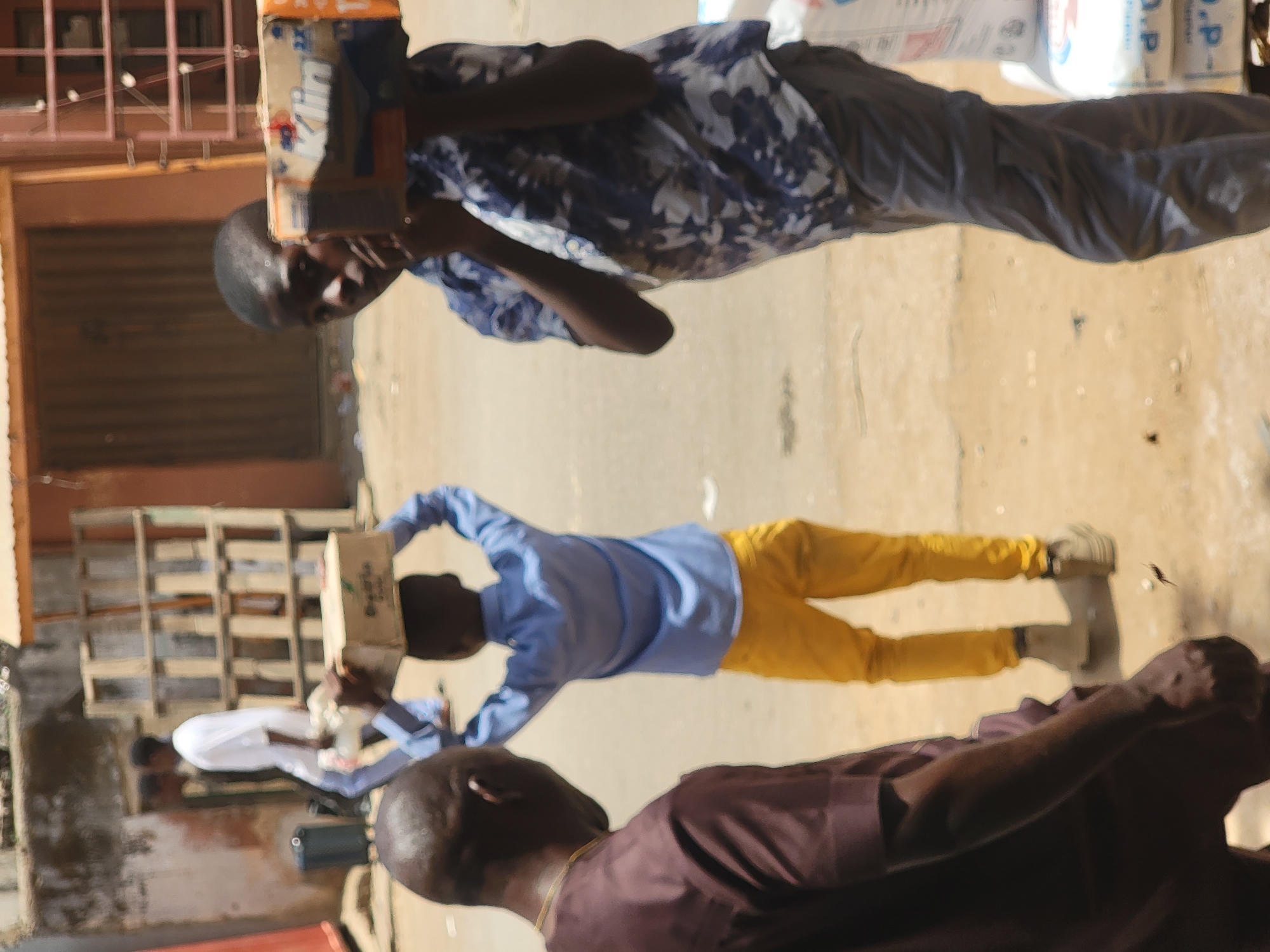 The image depicts an outdoor scene in Maiburiji, Nigeria, showing three men. One man is carrying a box on his head, dressed in a blue patterned shirt and gray pants. Another man, in a blue shirt and yellow pants, is also carrying a box on his head. A third man, wearing a brown shirt, is in the foreground. In the background, there is a building and several wooden shelves. Bags of products are also visible, adding context to the scene, presumably a market or storage area within the city.