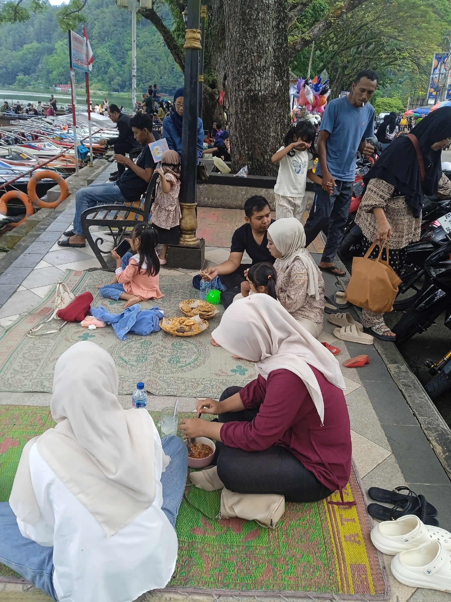 A group of people are sitting on mats on a paved area next to a body of water, likely a lake, in Nglingi, Indonesia. Some individuals are eating from bowls and plates, while others interact with each other. There are boats docked along the shoreline in the background. Some people are sitting on a bench near a lamppost. A father and daughter are holding hands and walking near a parking lot with several motorcycles.